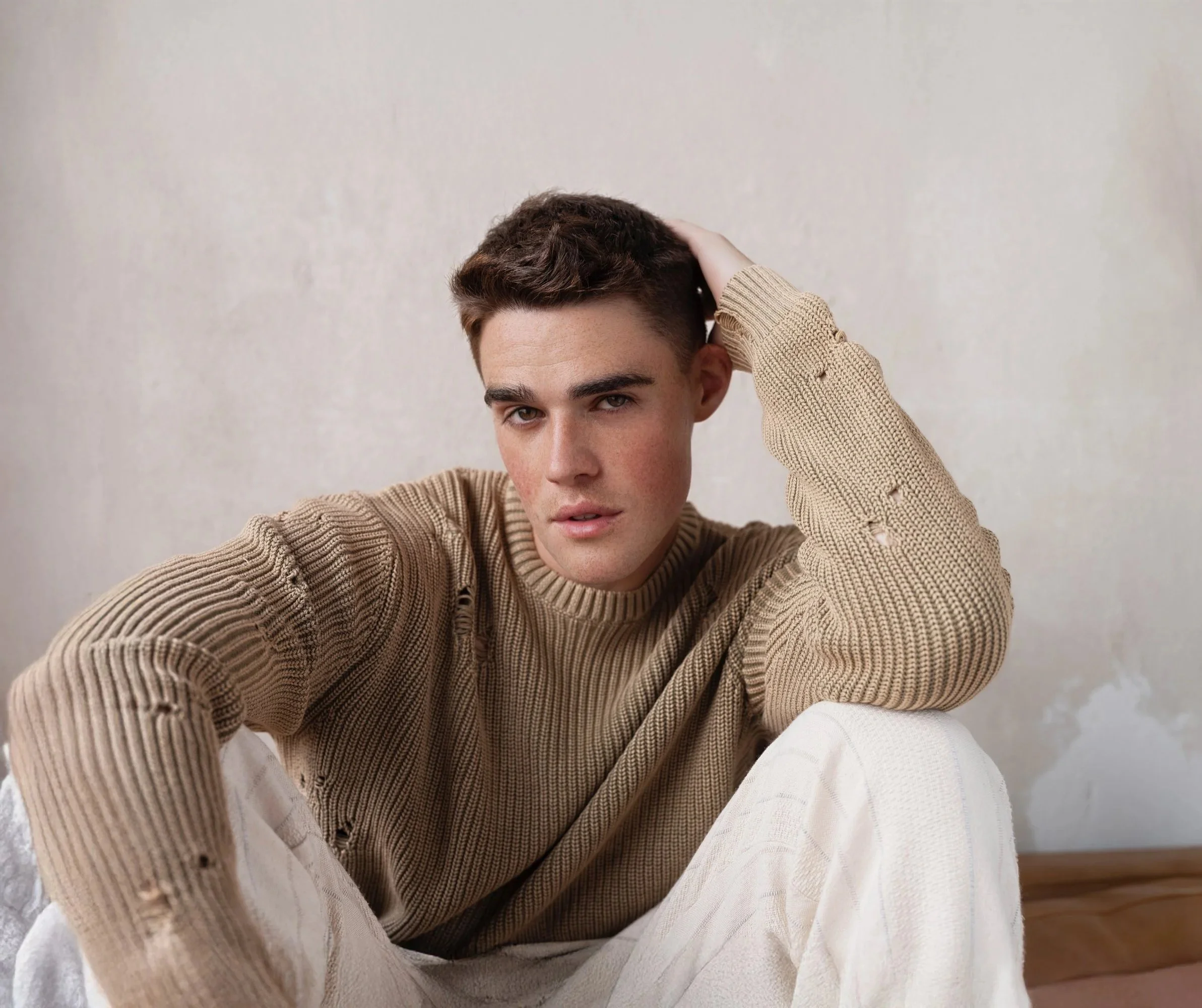 A young man with dark hair, wearing a distressed beige sweater and white pants, sitting on the floor with one arm resting on his knee and the other hand in his hair, in front of a plain wall.