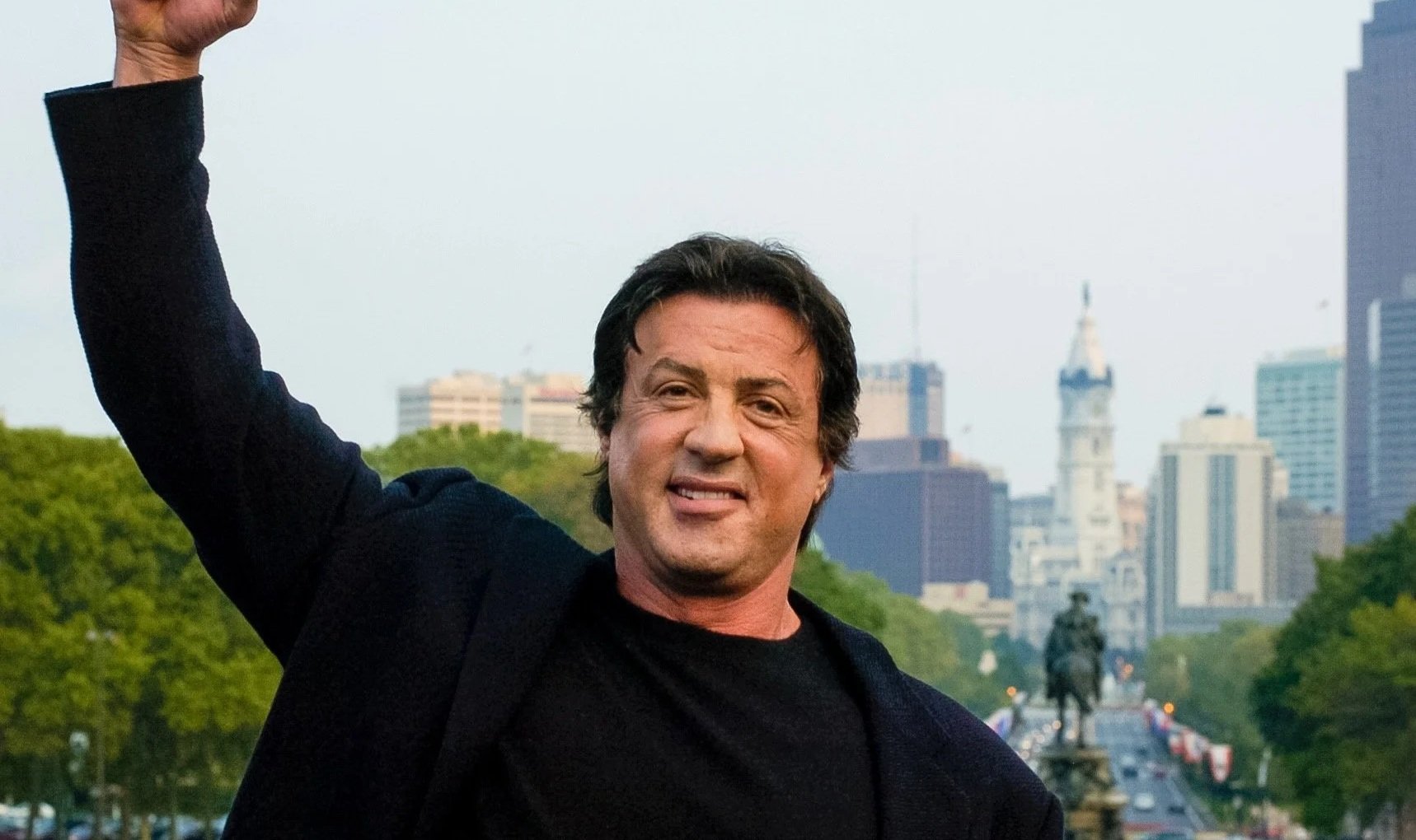 A man with dark hair wearing a black shirt and jacket, smiling and raising his arm, with a cityscape and statue in the background.