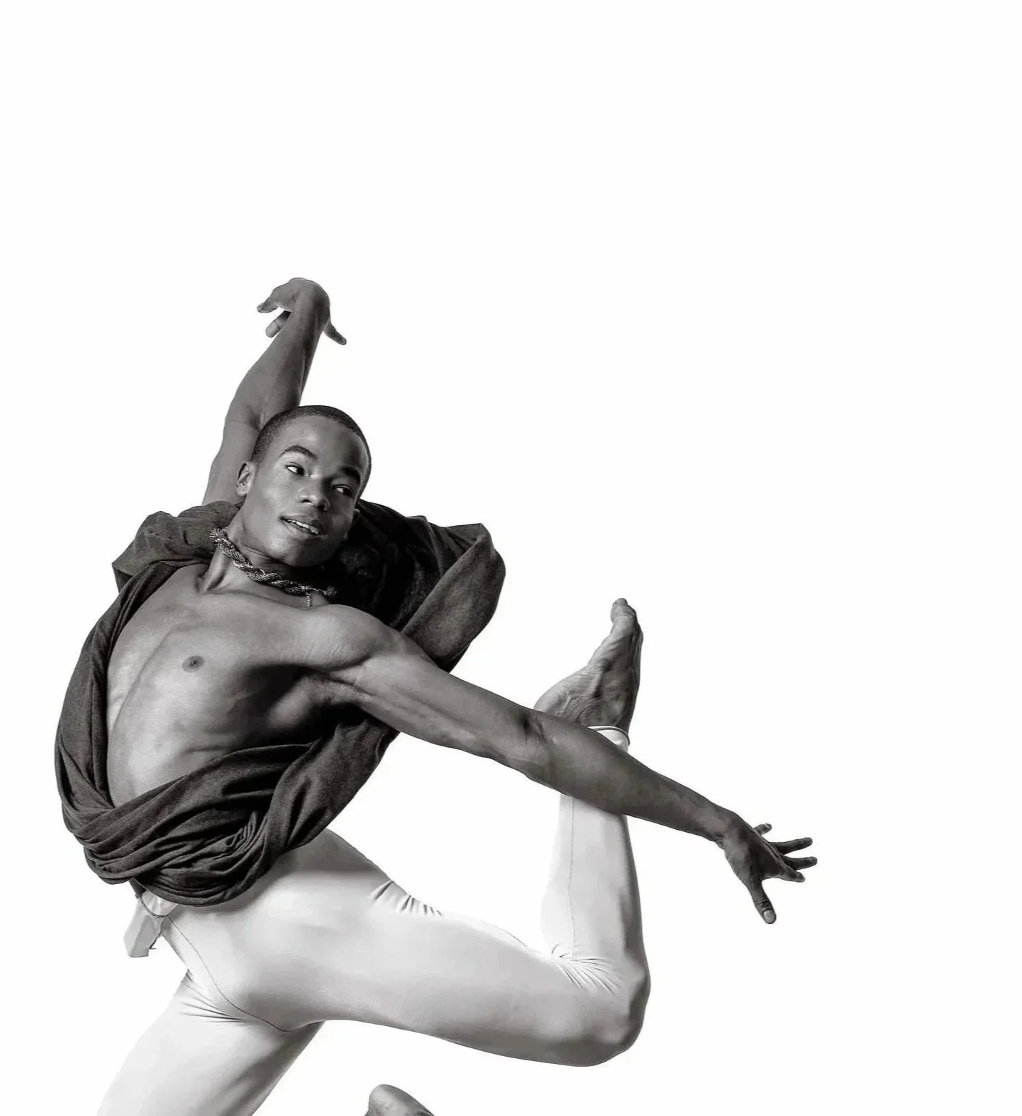 A black and white photo of a person ballet dancing with an expressive pose, wearing tight leggings and a draped top, against a plain background.