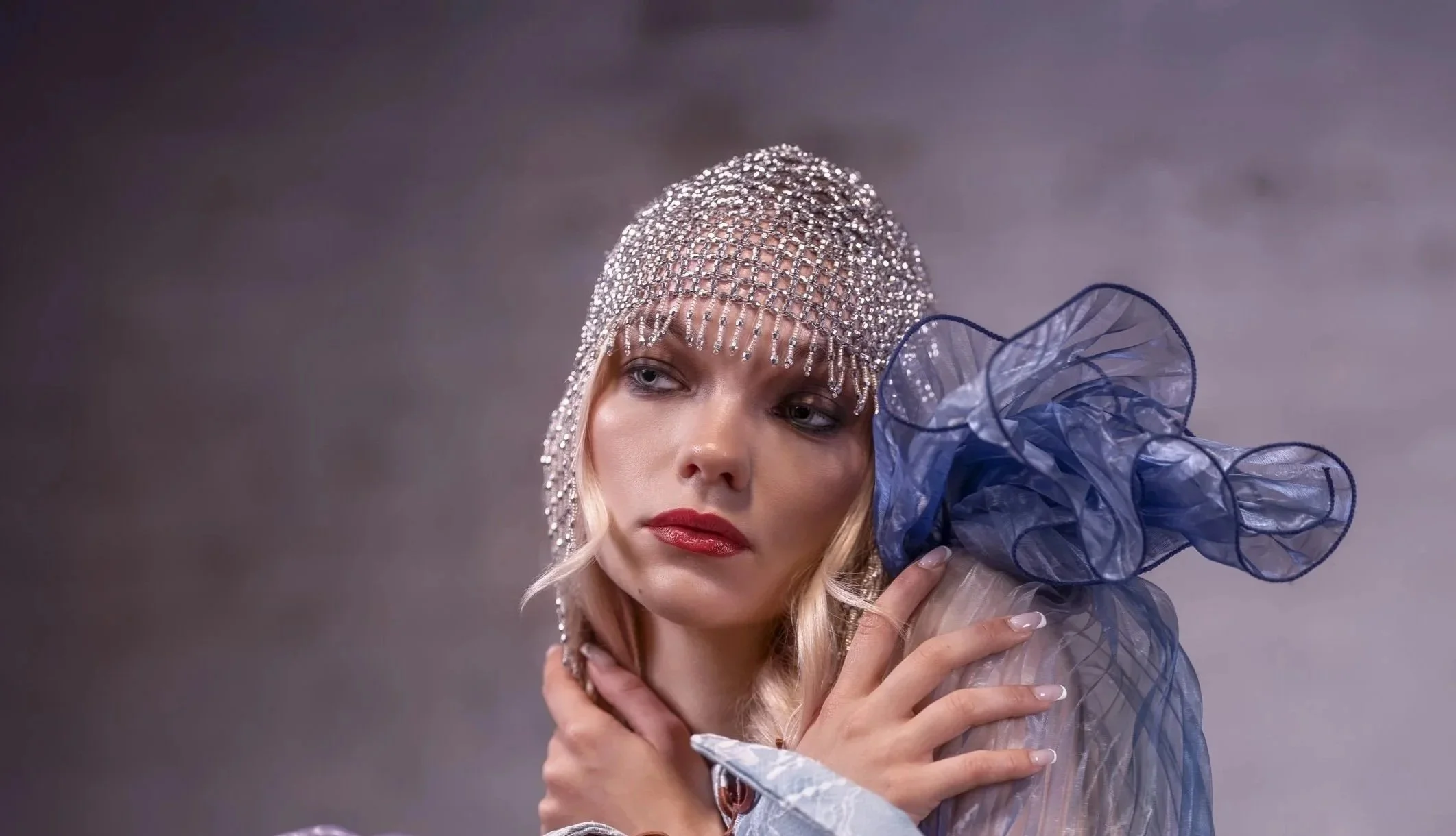 A woman with blonde hair wearing a rhinestone-covered headpiece and a sheer blue ruffled sleeve holds her hand to her neck, against a gray background.