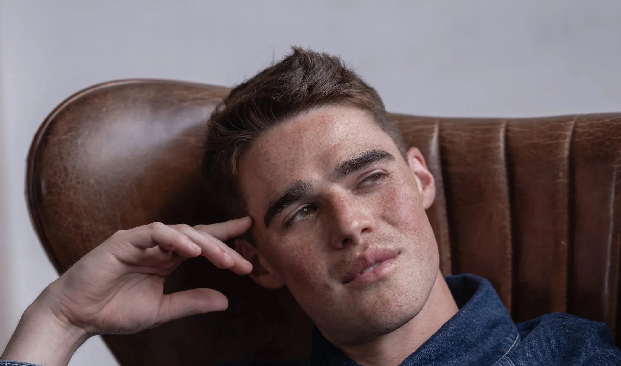 Close-up of a young man with brown hair and freckles, reclining on a wooden chair, looking contemplative.