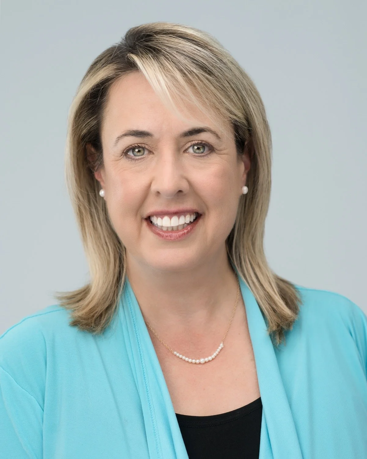 A professional woman with shoulder-length blonde hair, wearing a blue blazer, black top, pearl necklace, and matching pearl earrings, smiling against a light gray background.
