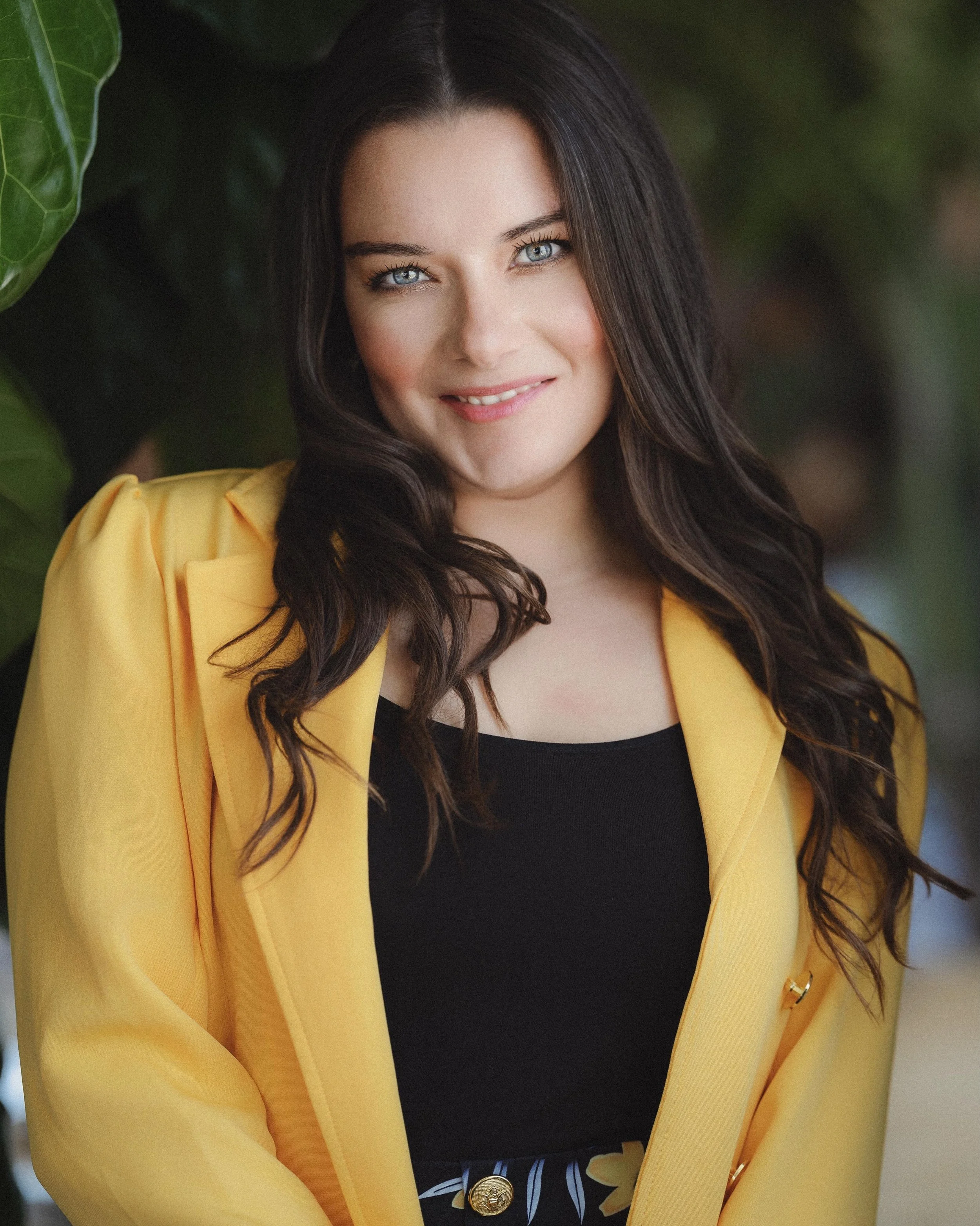 A young woman with blue eyes, long dark hair, and fair skin, wearing a yellow blazer over a black top, smiling at the camera.