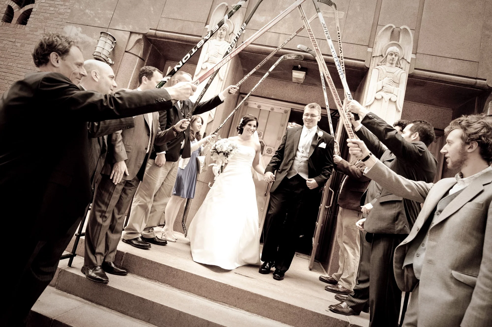 Bride and groom walking out of a building, surrounded by friends and family holding hockey sticks in celebration, during a wedding ceremony.