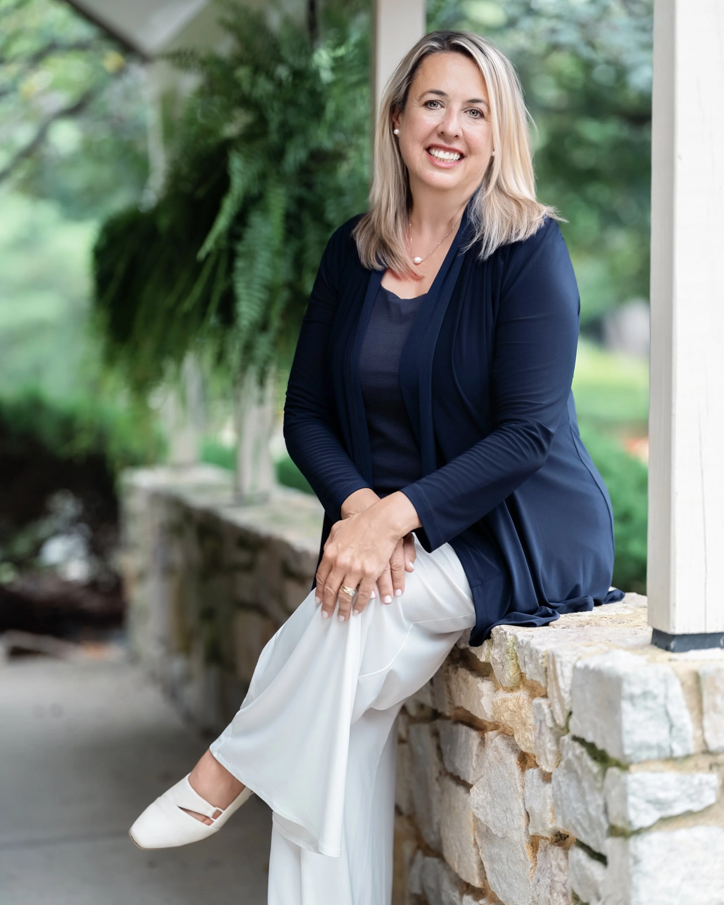 A woman with blonde hair sitting on a stone wall outdoors, wearing a navy blazer, navy top, and white pants, smiling at the camera.
