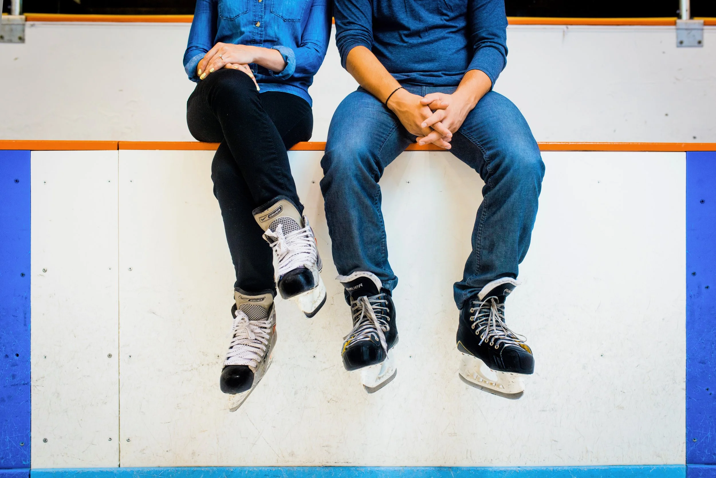 Two people sitting on an ice rink ledge wearing ice skates, with their hands resting on their laps, one in black jeans and the other in blue jeans.