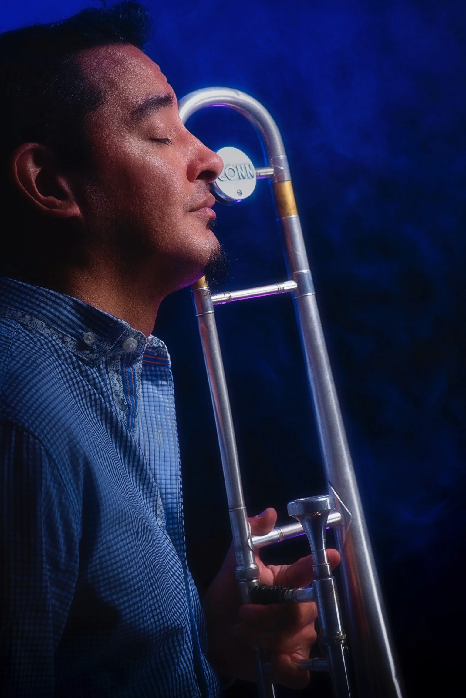 Man with closed eyes holding a trombone against his face, wearing a checkered shirt, with a dark blue background.