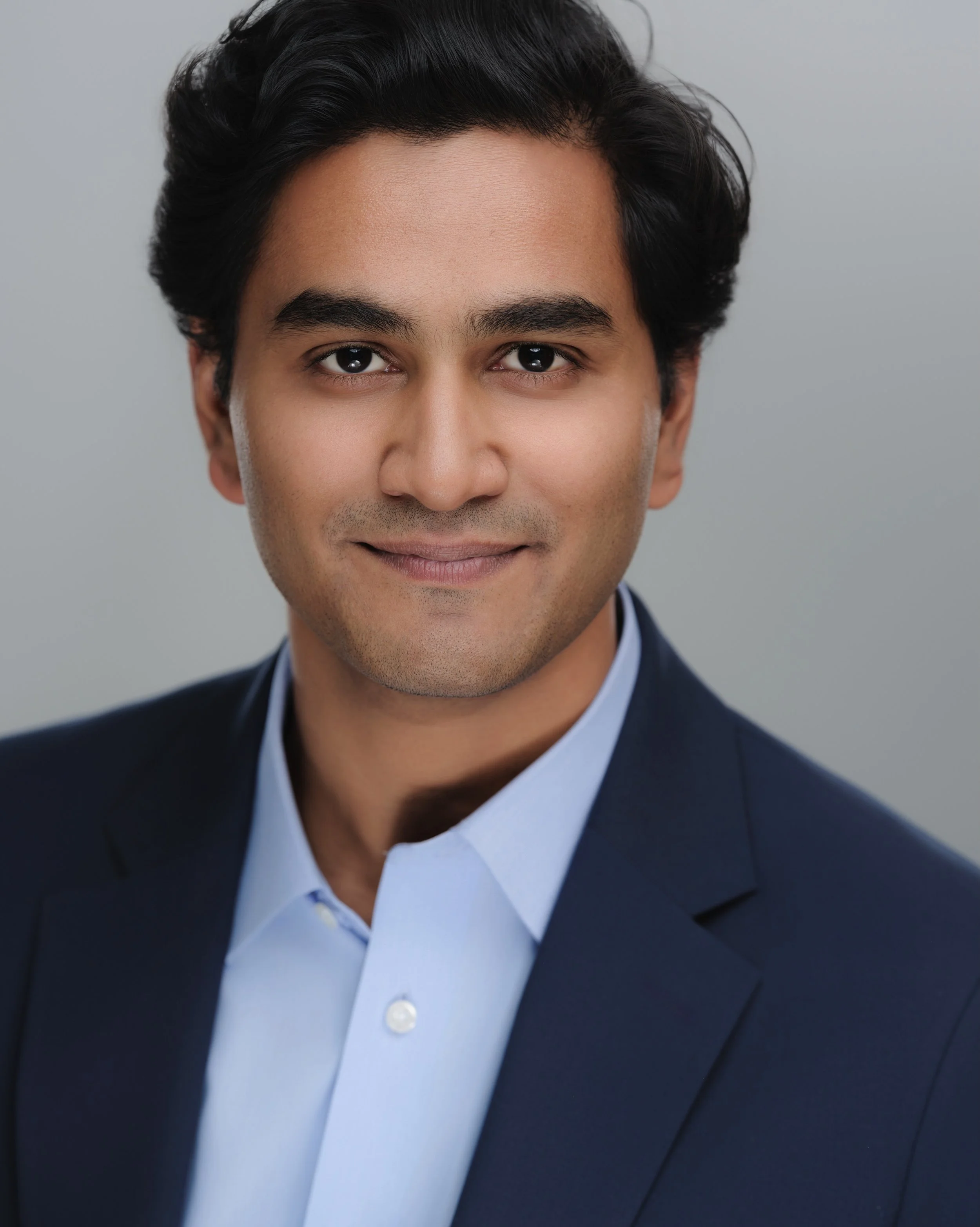 Headshot of a young man with dark hair, wearing a navy blazer and light blue shirt, smiling softly against a neutral background.