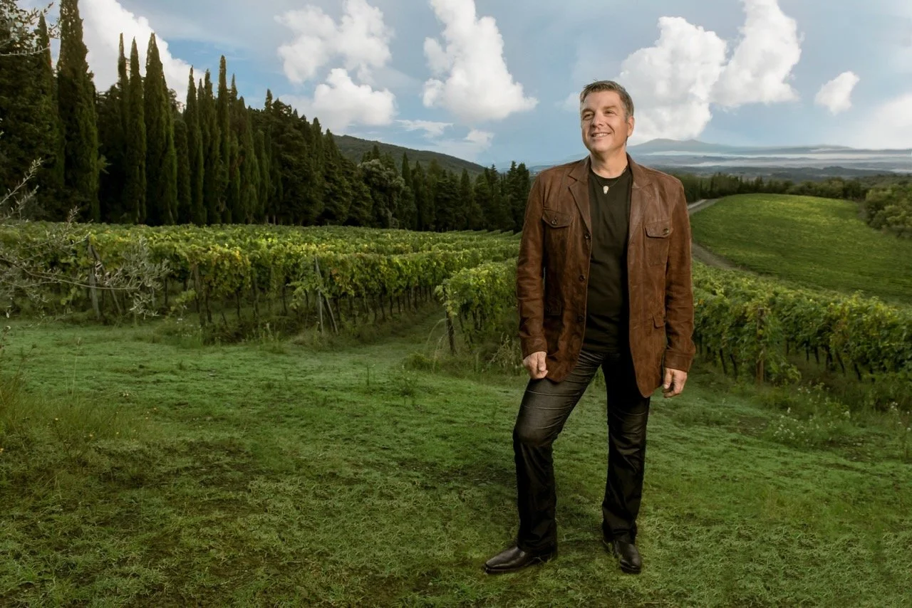 A man stands in a vineyard with lush green grapevines, tall cypress trees, and rolling hills under a partly cloudy sky.