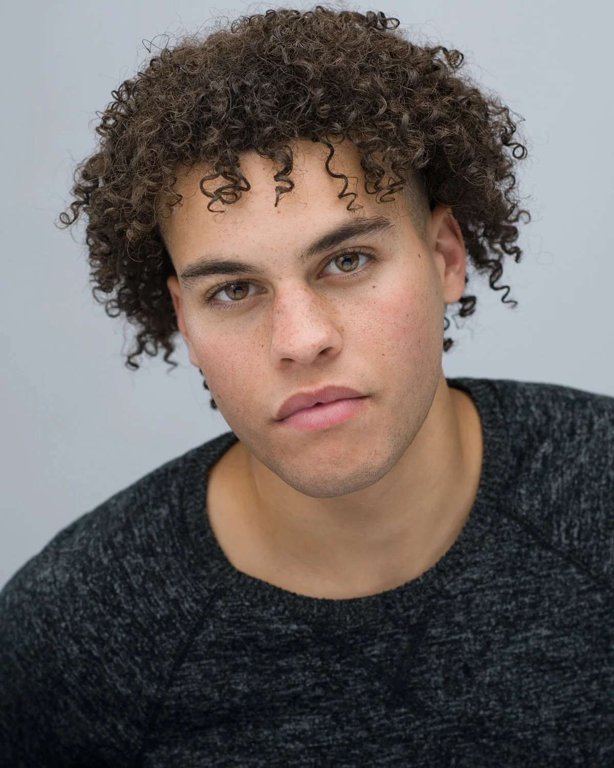 Close-up portrait of a young man with curly hair, hazel eyes, and light skin, wearing a dark gray sweater, looking directly at the camera with a neutral expression.