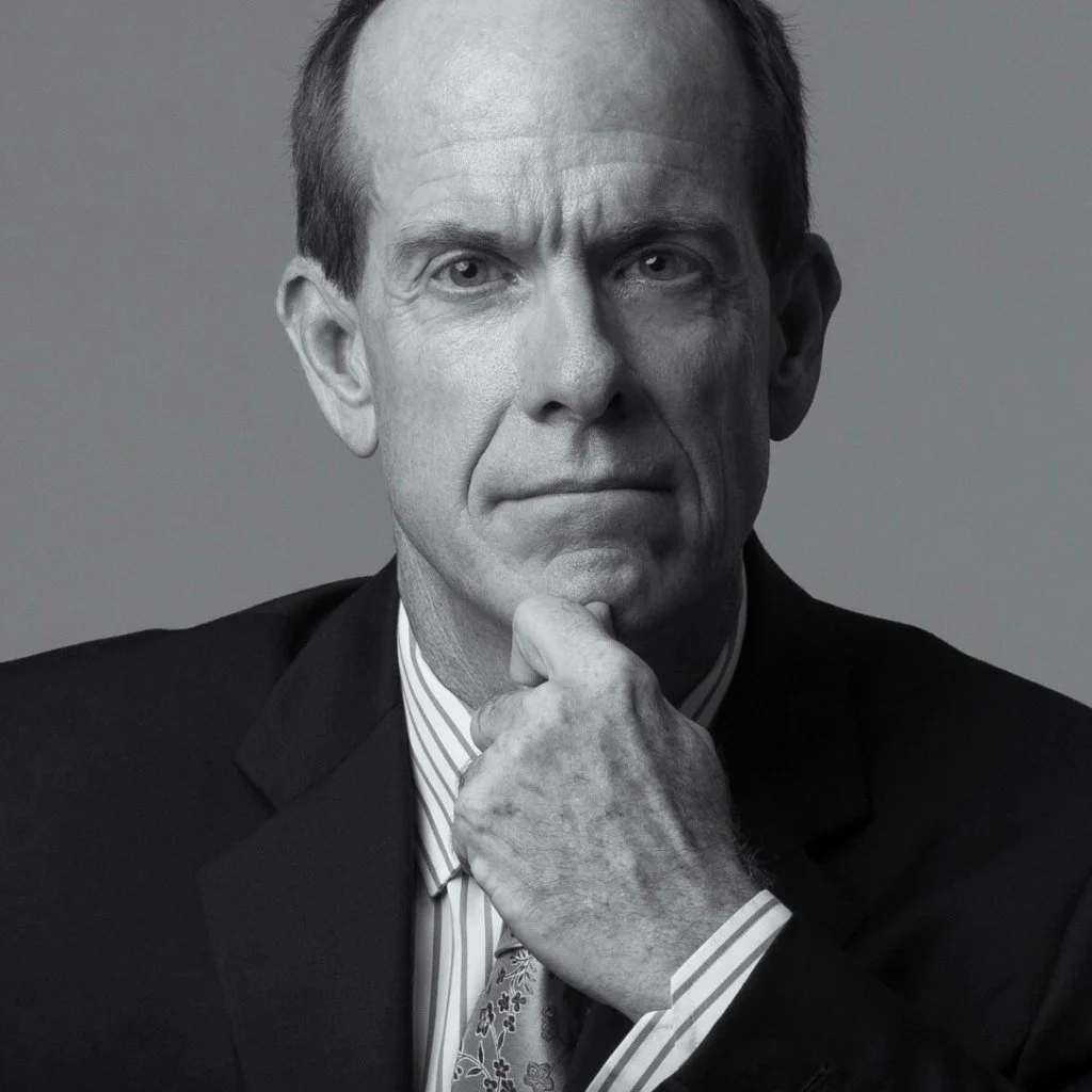 Black and white portrait of a middle-aged man with short hair, wearing a suit and tie, holding his chin with his hand, looking directly at the camera.
