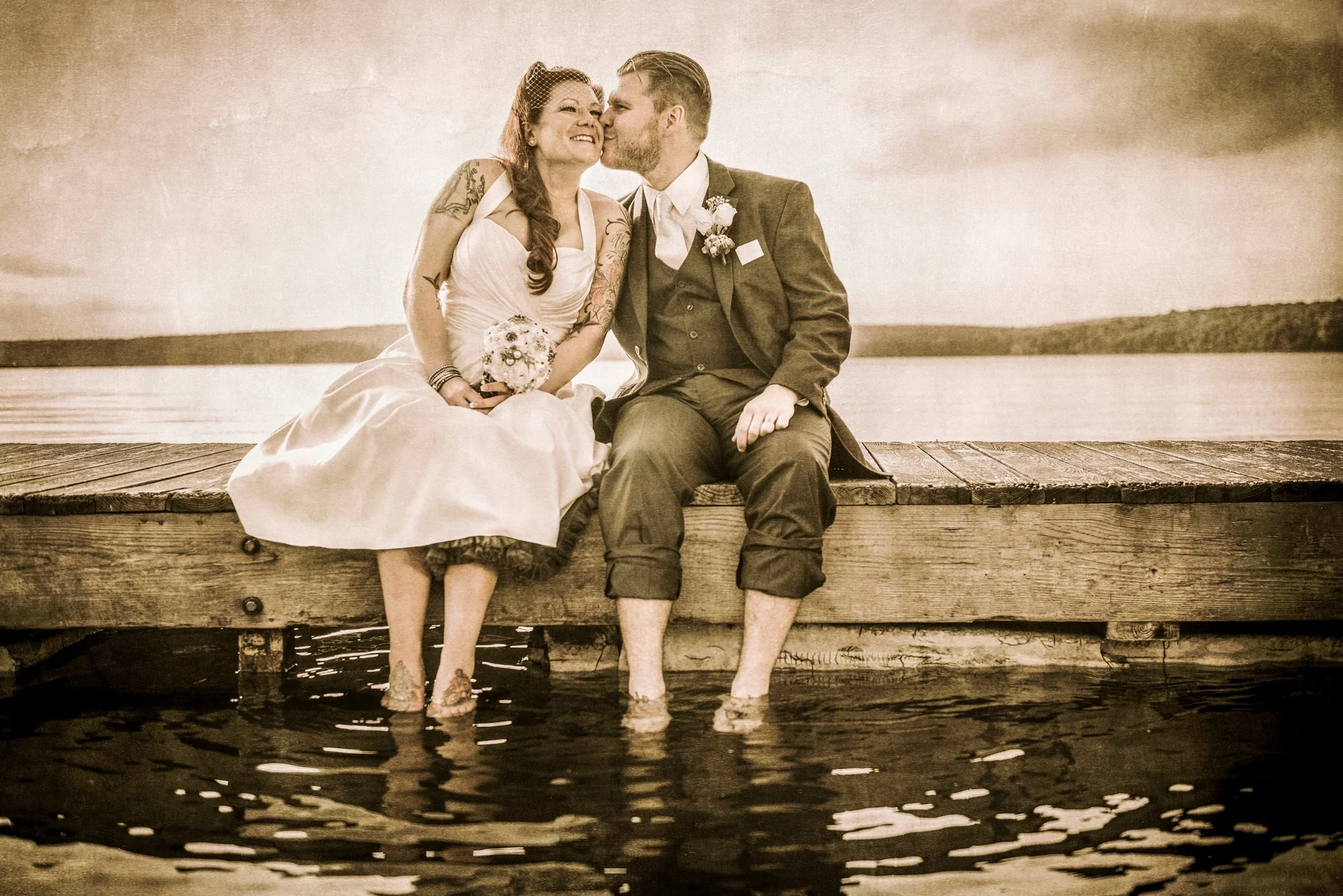 A couple dressed in wedding attire sitting on a wooden dock over water, with the bride holding a small bouquet. The bride and groom are kissing, with their feet submerged in the water, and a scenic landscape in the background.