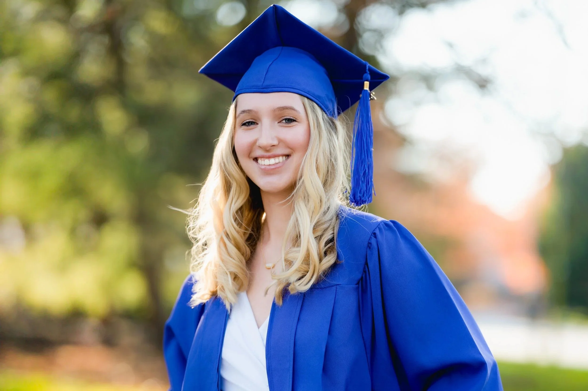 Smiling young woman in a blue graduation cap and gown outdoors with trees and sunlight in the background.