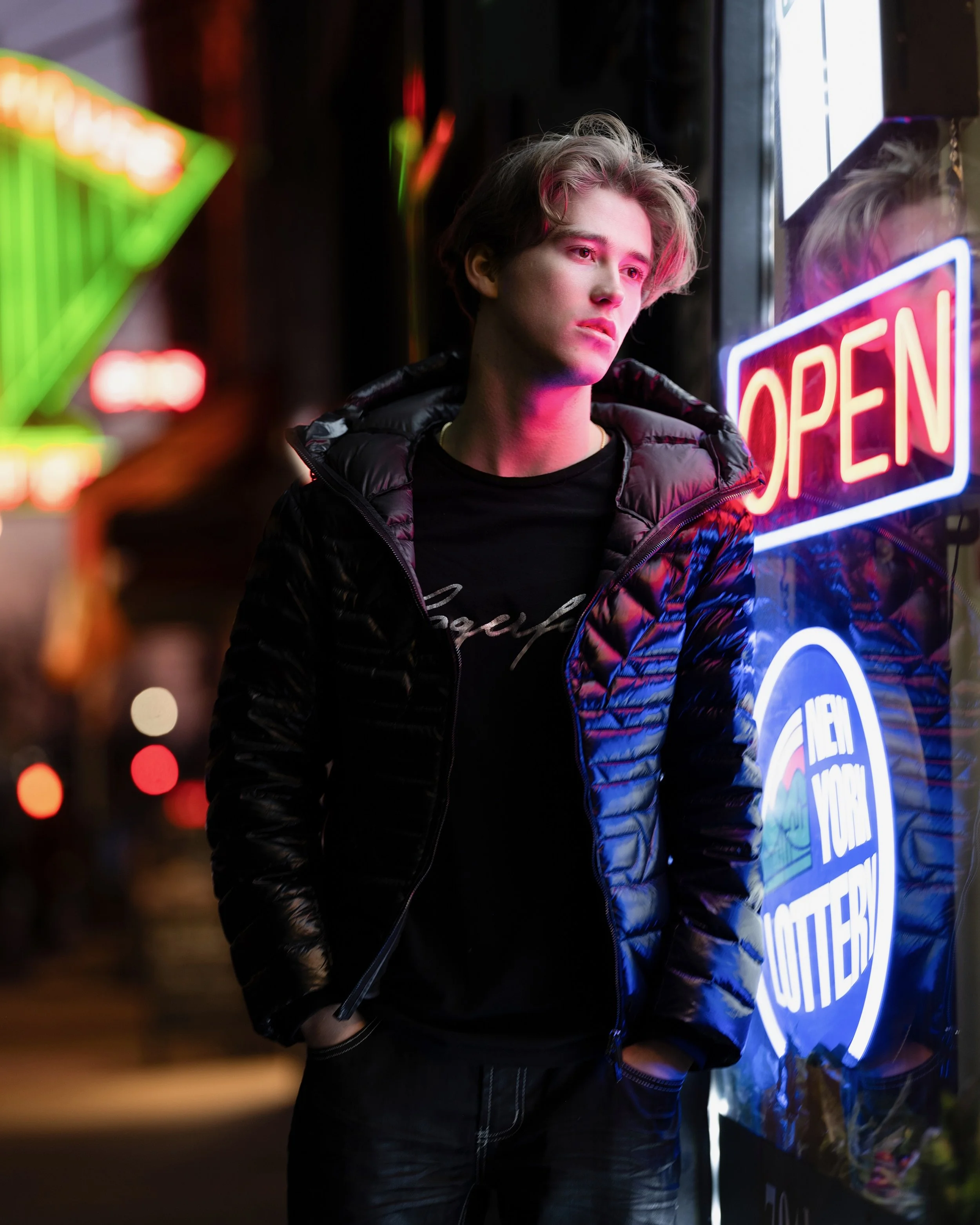 Young man standing outside at night near a neon 'OPEN' sign, wearing a black jacket and black shirt.