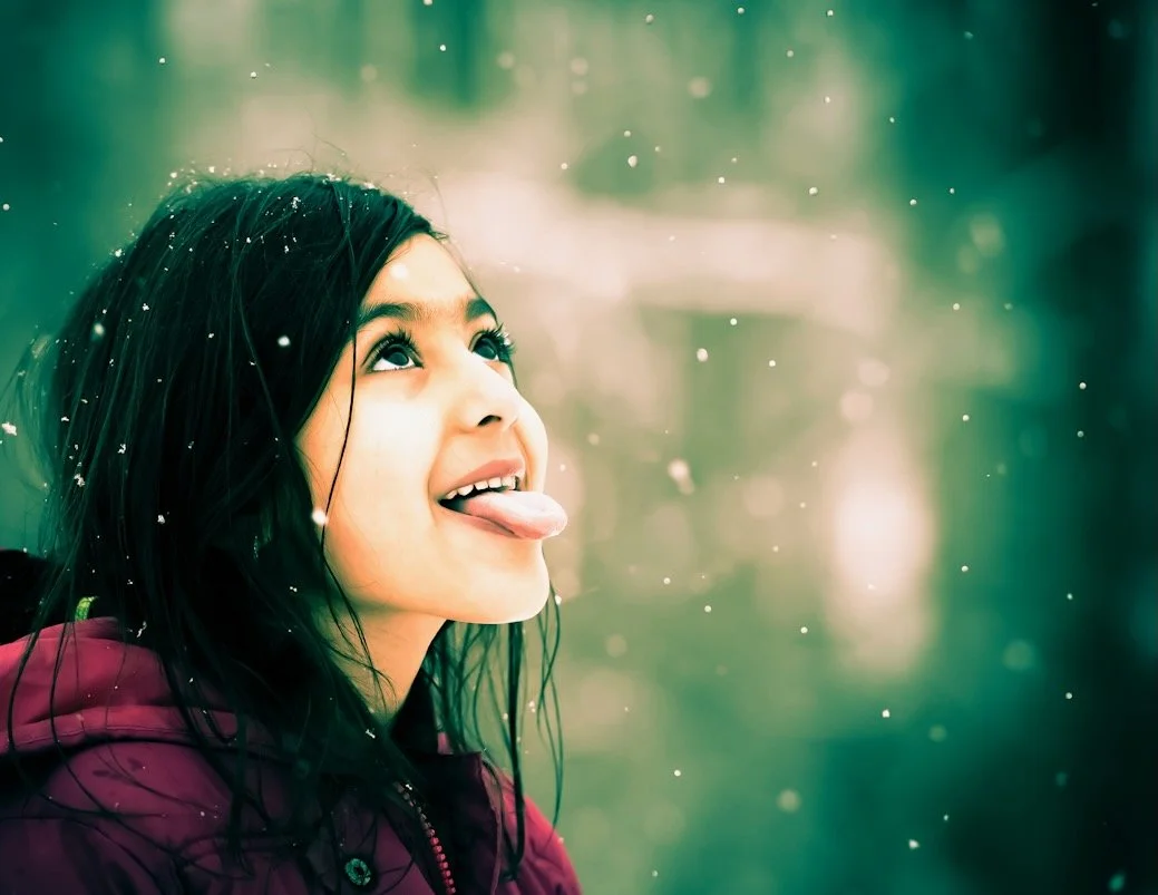 A young girl with dark hair and a maroon jacket sticking out her tongue and looking upward in a snowy outdoor setting.