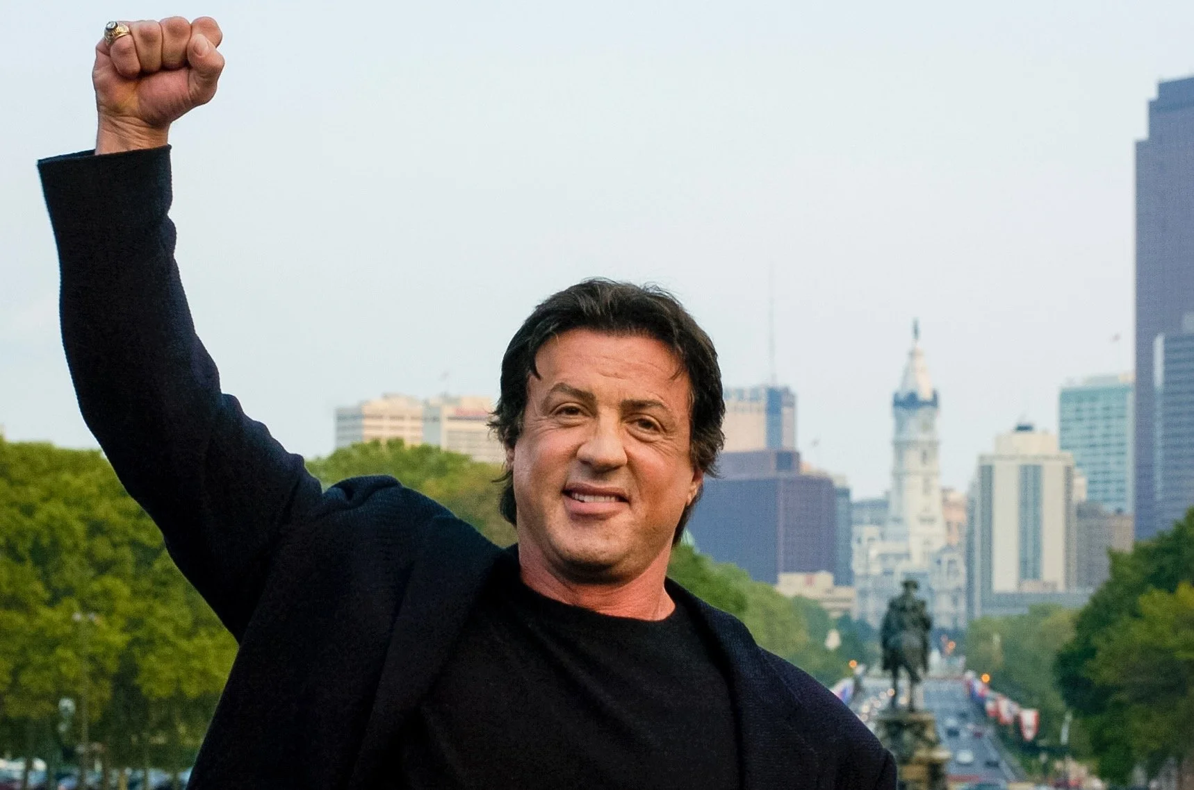 A man with dark hair, wearing a black jacket and black shirt, raising his right fist in the air outdoors with city buildings and trees in the background.