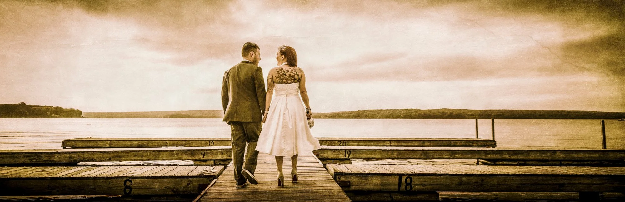 A couple in wedding attire holding hands and walking on a dock near a lake, with an overcast sky.