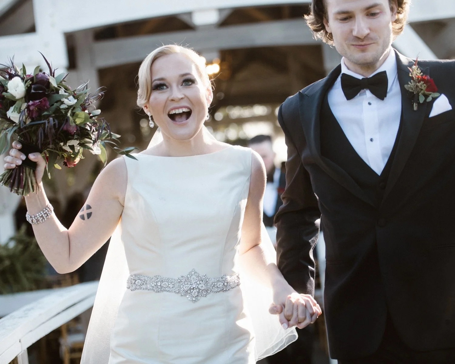 A joyful bride in a white wedding dress holding a bouquet, walking hand in hand with a groom in a black tuxedo with a bow tie, during a wedding ceremony outdoors.