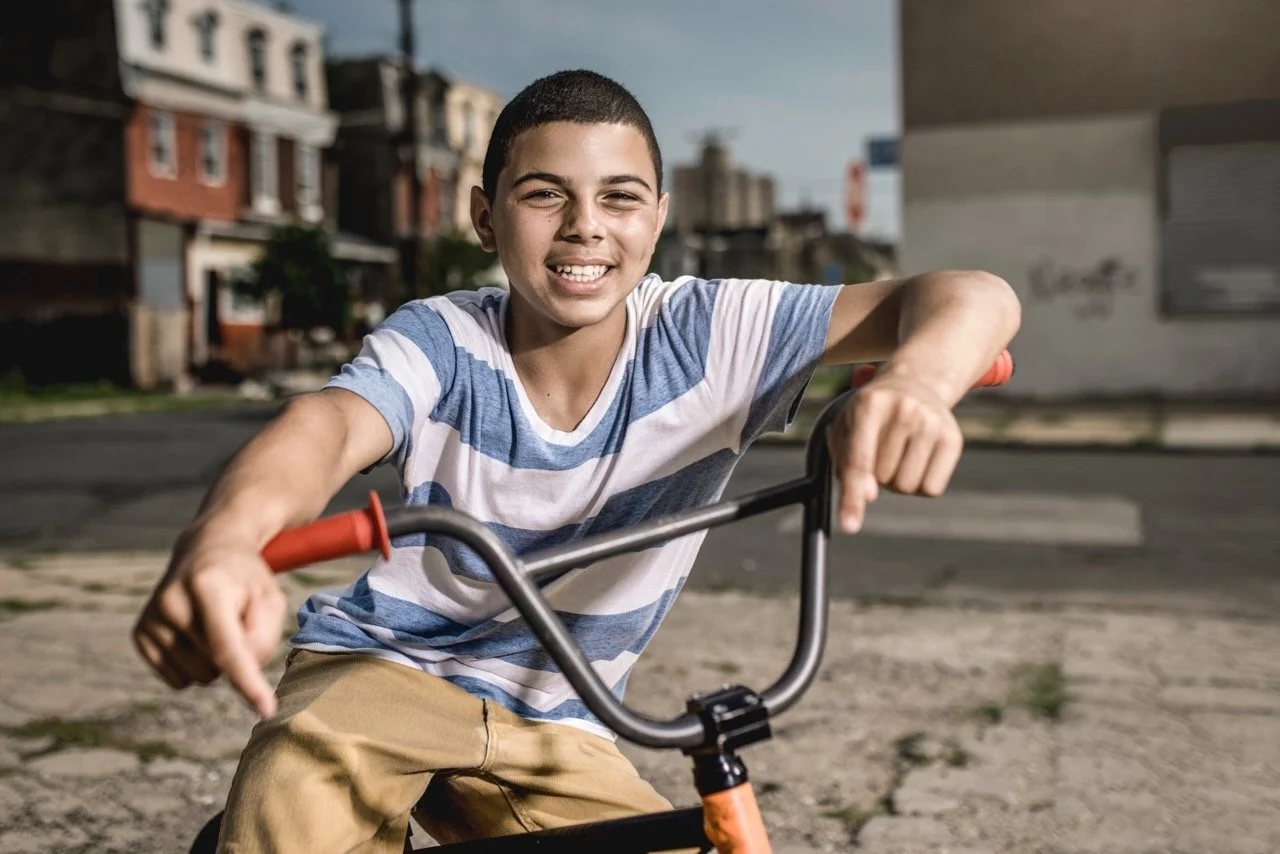 A boy smiling while sitting on a bicycle outdoors in a neighborhood.