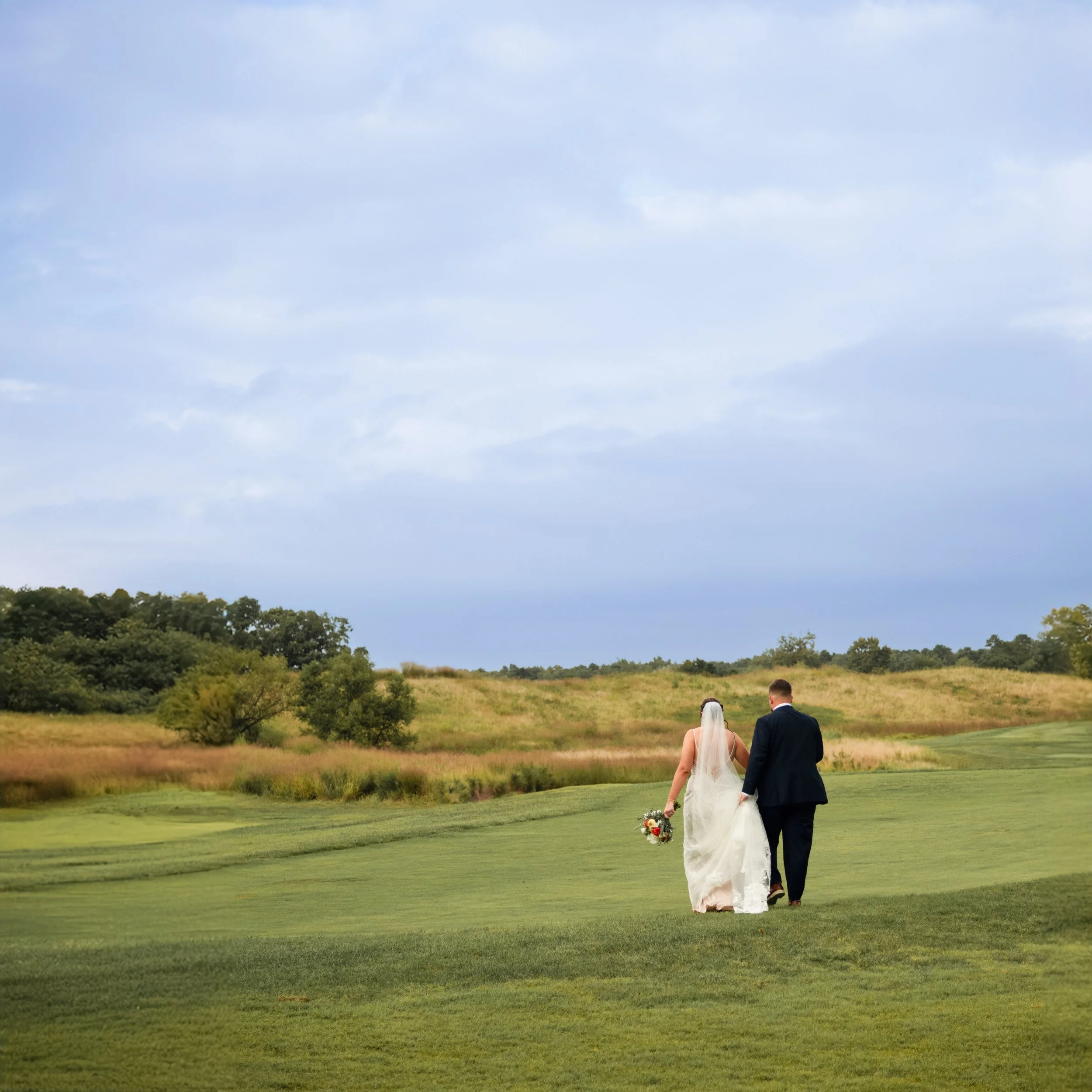 Bride and groom walking hand in hand across a grassy field, with the bride holding a bouquet, under a cloudy sky.