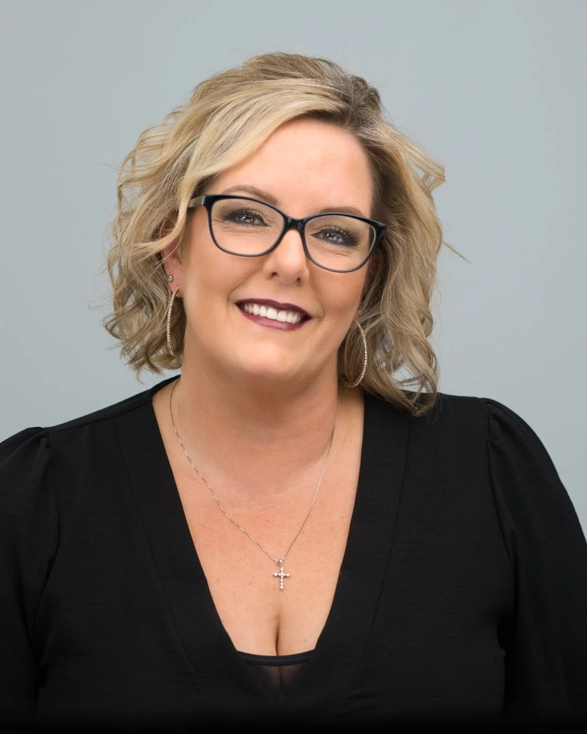 Smiling woman with blonde hair, black glasses, hoop earrings, and a cross necklace, wearing a black top.