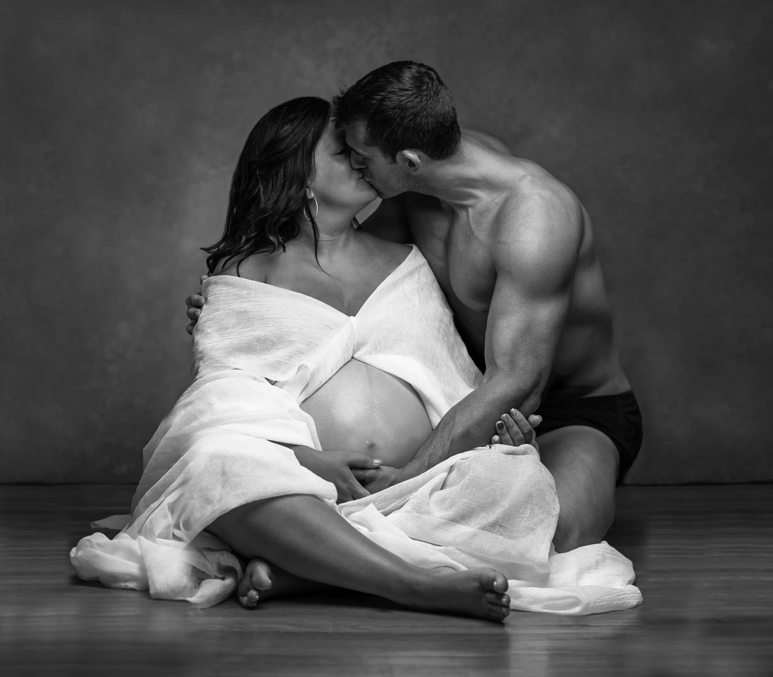 A pregnant woman and a man are kissing, sitting on a floor with a plain backdrop; she is wearing a white dress, he is shirtless and wearing black shorts.