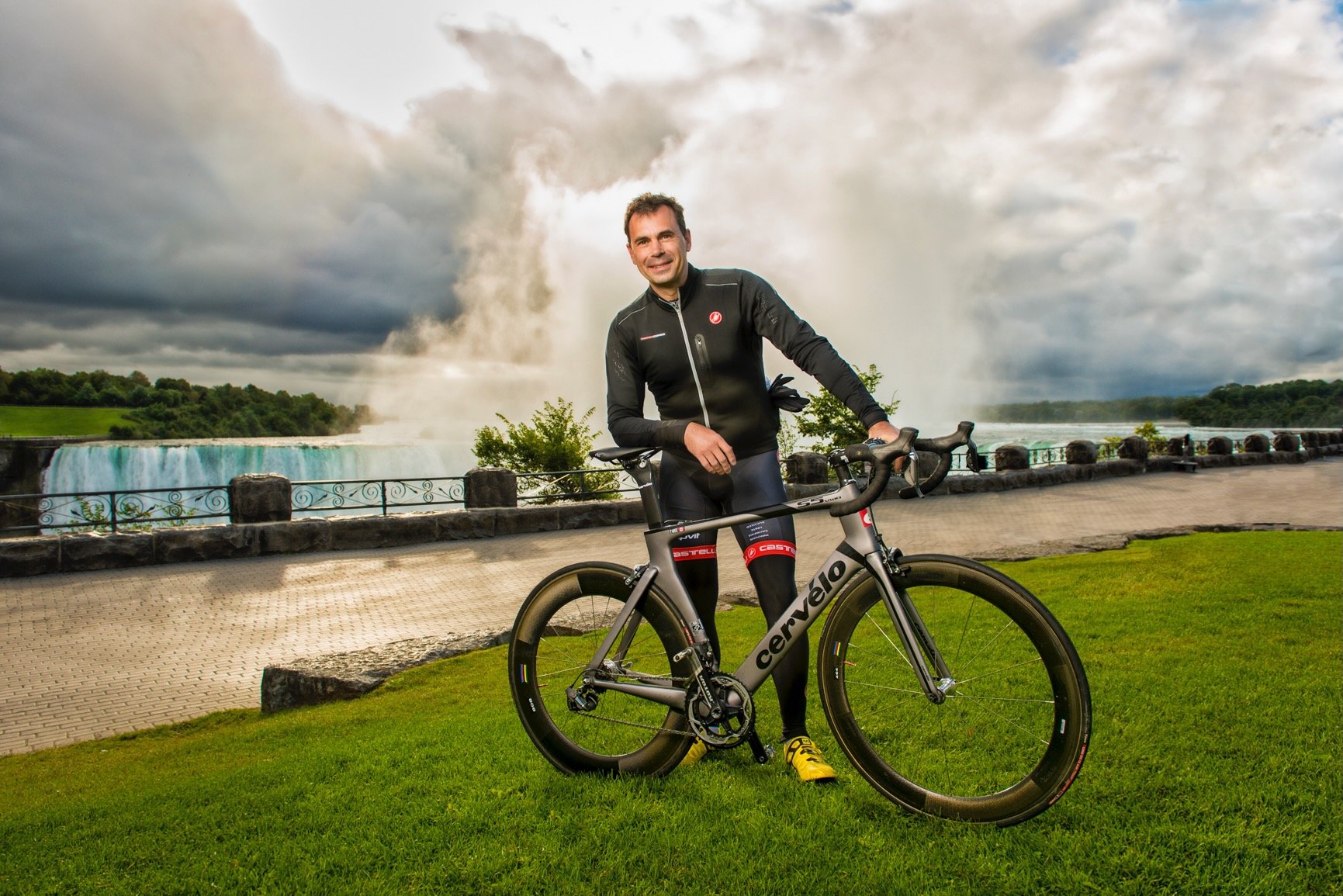Man in black cycling gear standing with a grey carbon bike in front of Niagara Falls during daytime.