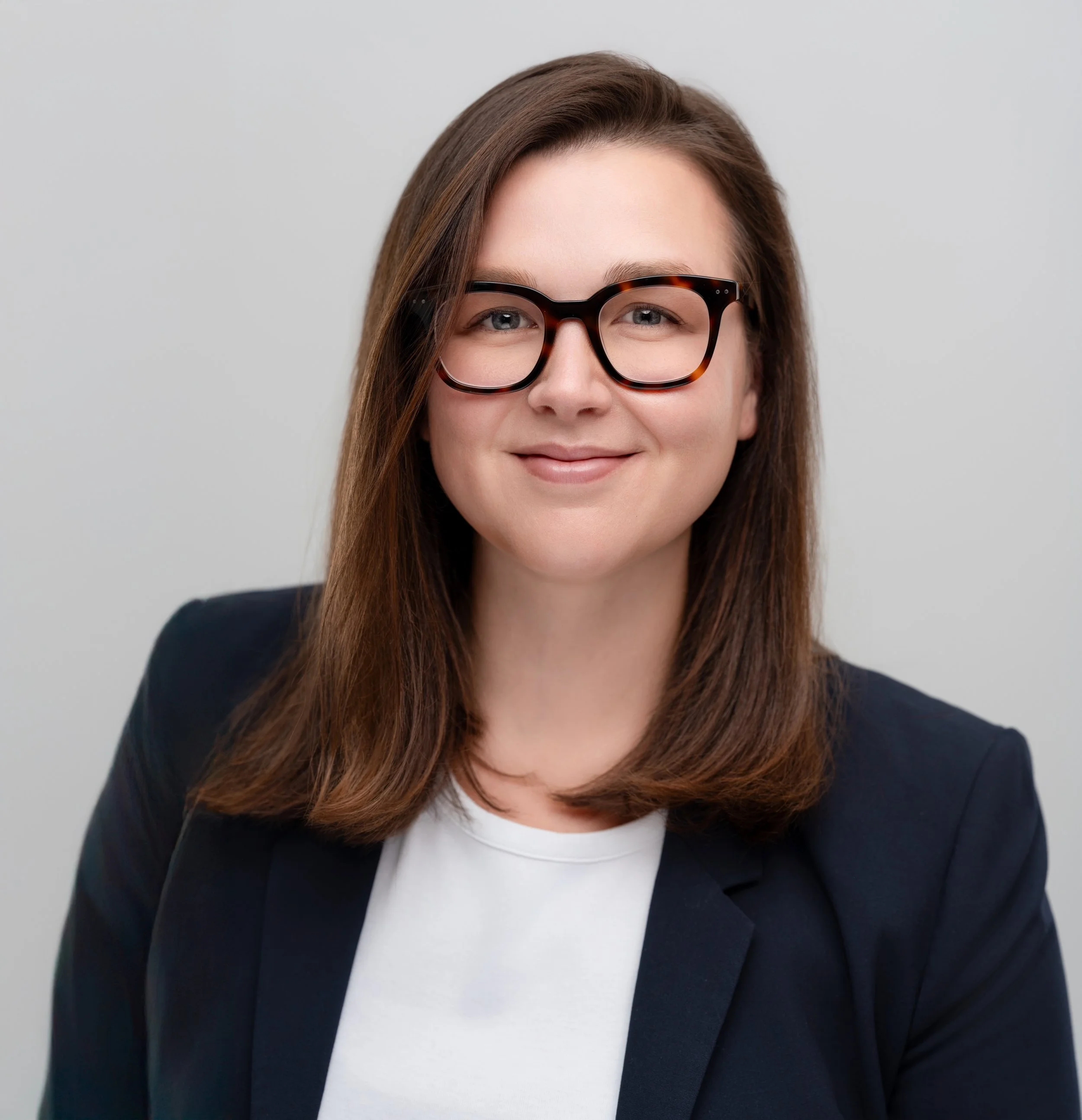 A woman with shoulder-length brown hair wearing glasses, a white shirt, and a dark blazer, smiling at the camera.
