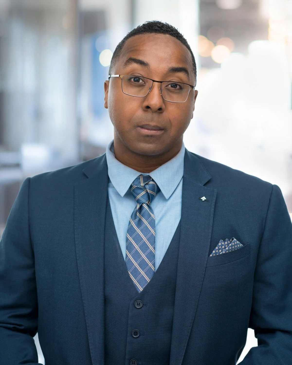 A man in a blue suit, light blue shirt, and patterned tie, wearing glasses, standing in an indoor setting with blurred background.