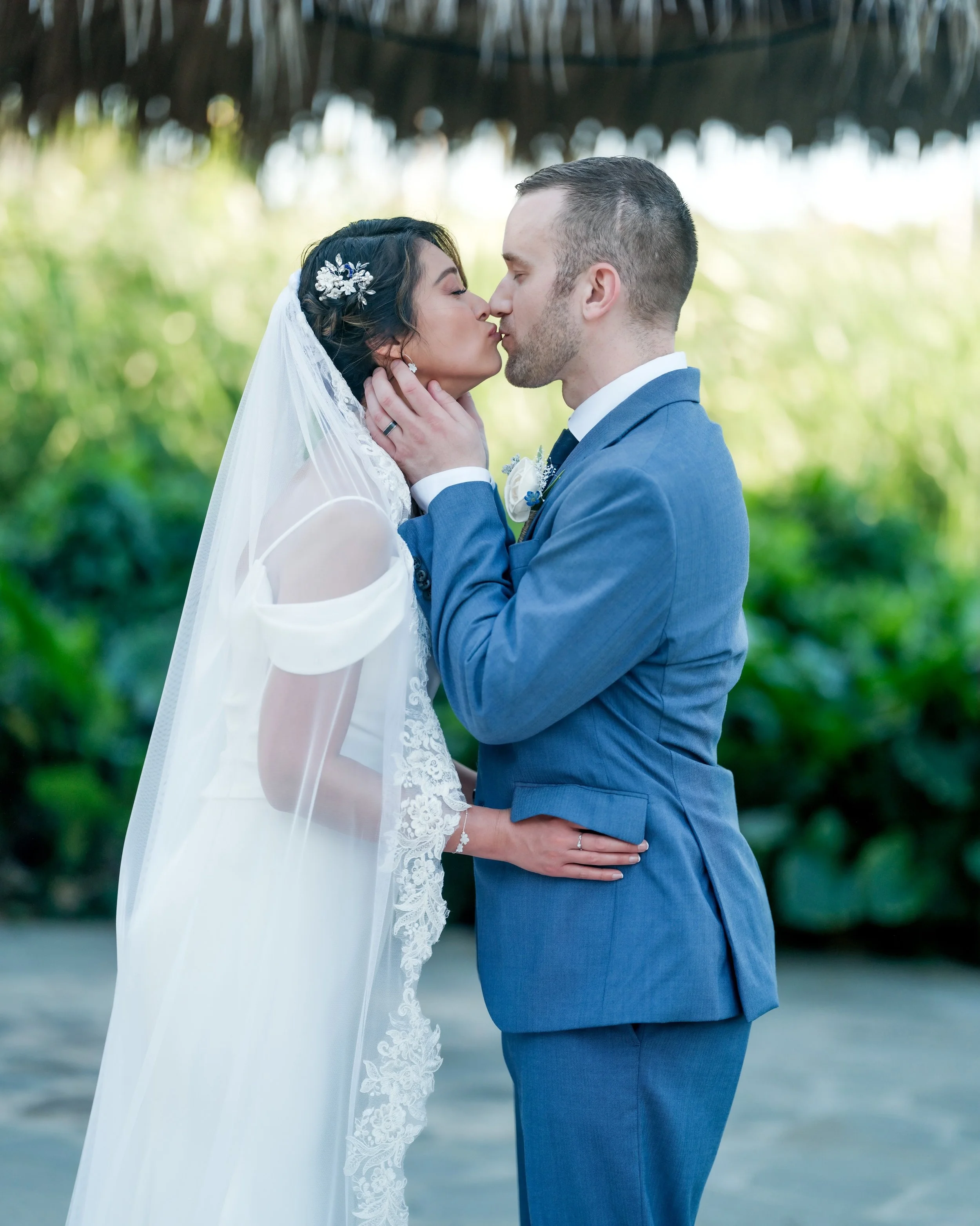 A bride and groom are about to kiss during their wedding ceremony, standing outdoors under a bamboo roof, with green foliage in the background.
