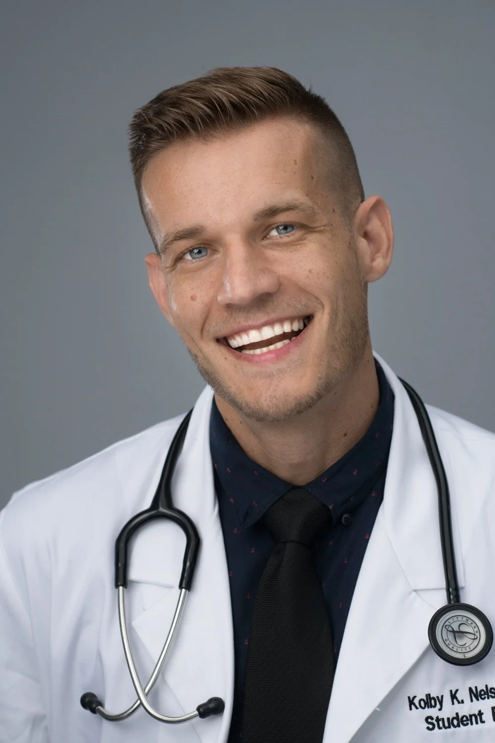 A young male doctor or medical student smiling, wearing a white lab coat with a stethoscope around his neck, against a plain gray background.