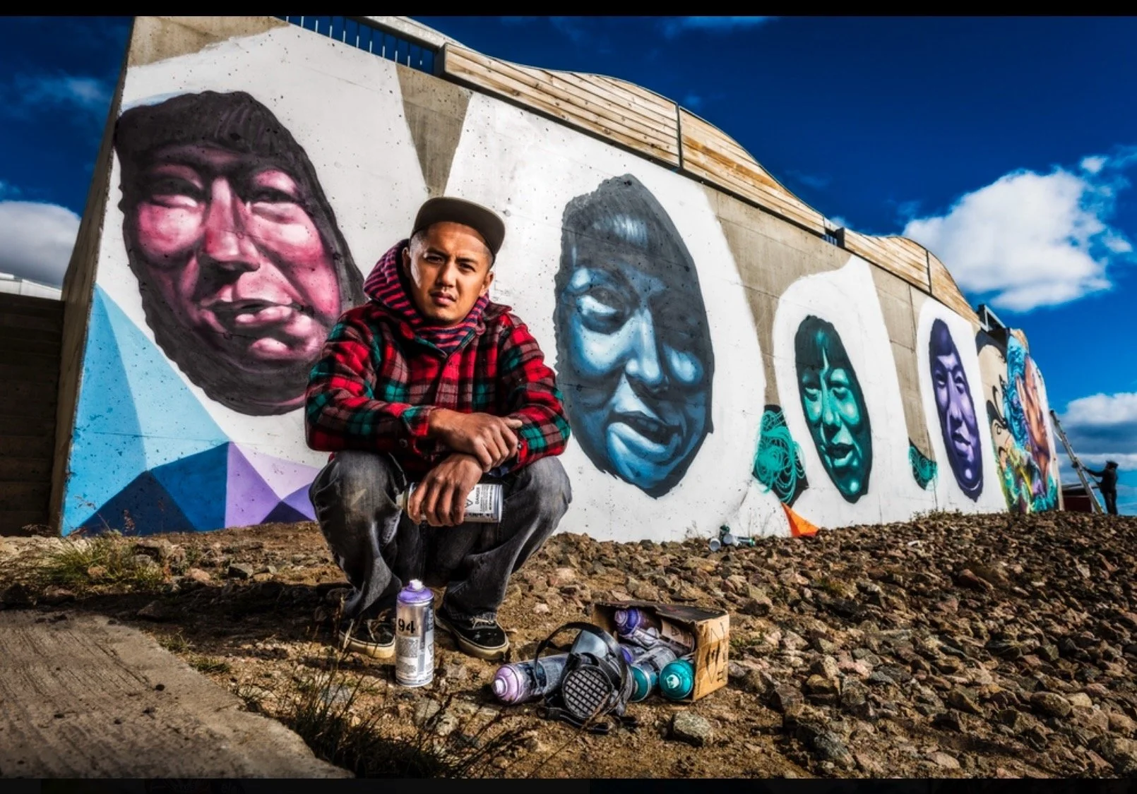 A young man squatting in front of a colorful mural of large, realistic portraits painted on a concrete wall. The mural features multiple faces, each in different shades of blue, purple, and pink. The man is wearing a red plaid jacket, a gray cap, and