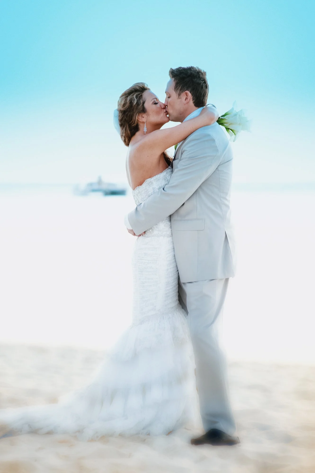 A newlywed couple kissing on the beach, with the bride in a white wedding dress and the groom in a light gray suit, holding a bouquet of white flowers.