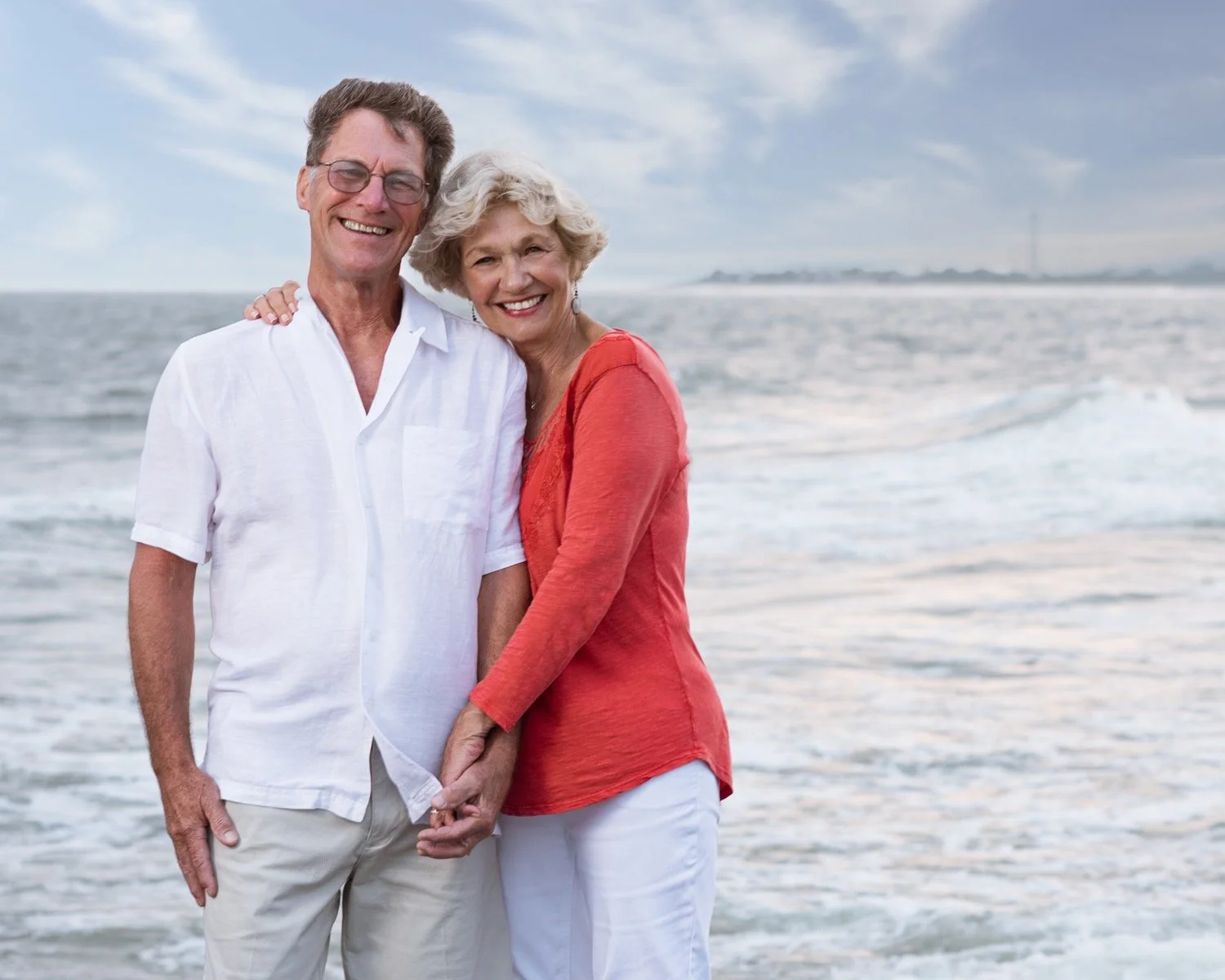 A smiling elderly couple holding hands on the beach with the ocean and cloudy sky in the background.