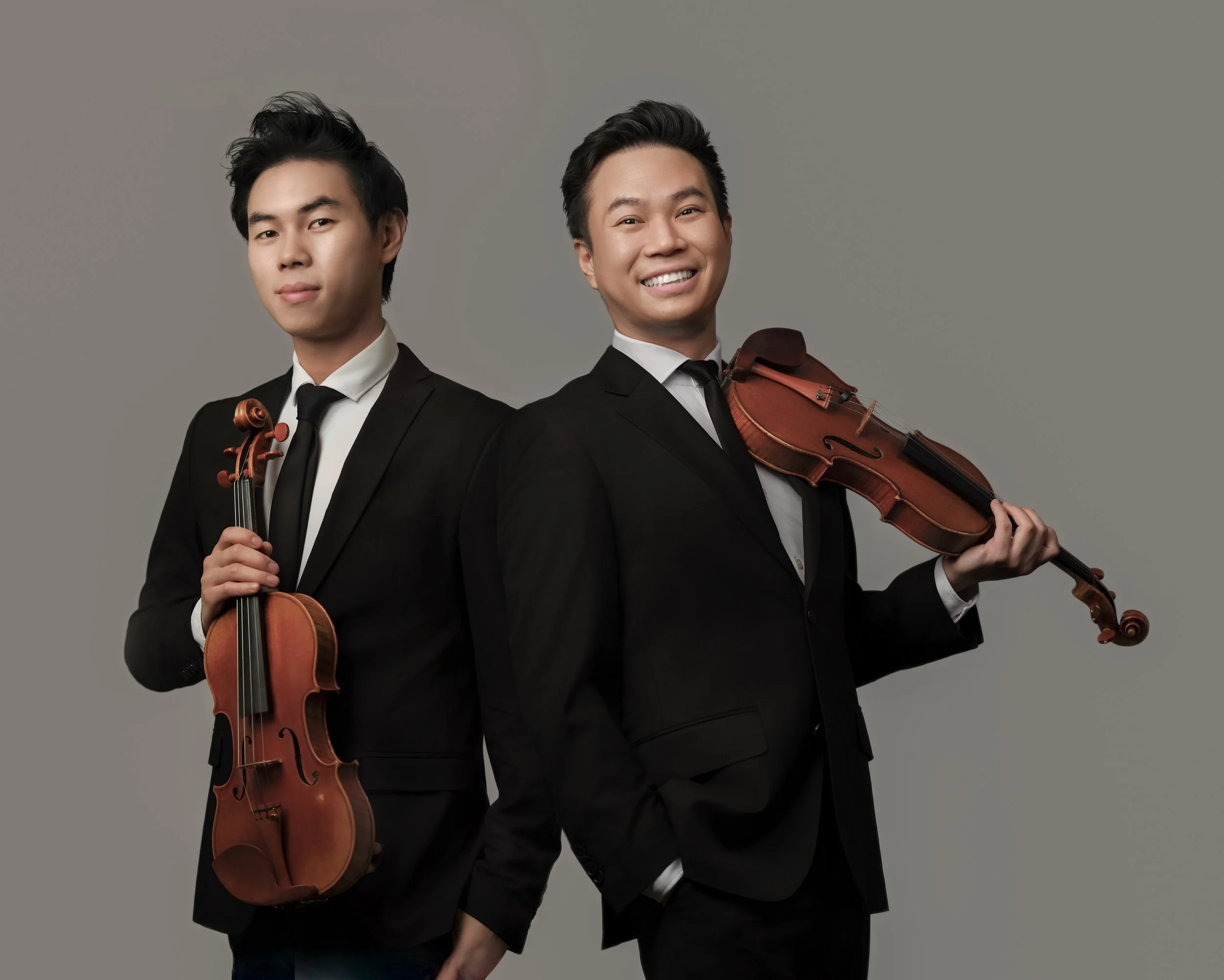 Two men in black suits holding violins, standing back-to-back against a grey background, one smiling and the other with a neutral expression.
