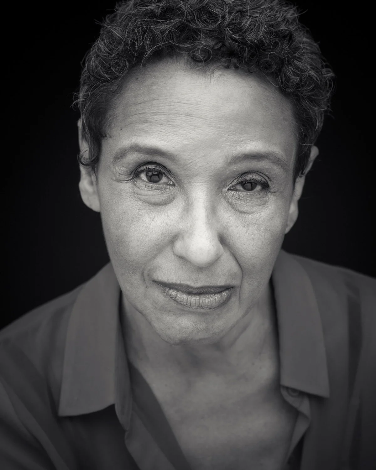 Black and white close-up portrait of a middle-aged woman with short, curly hair, wearing a collared shirt, looking into the camera against a dark background.