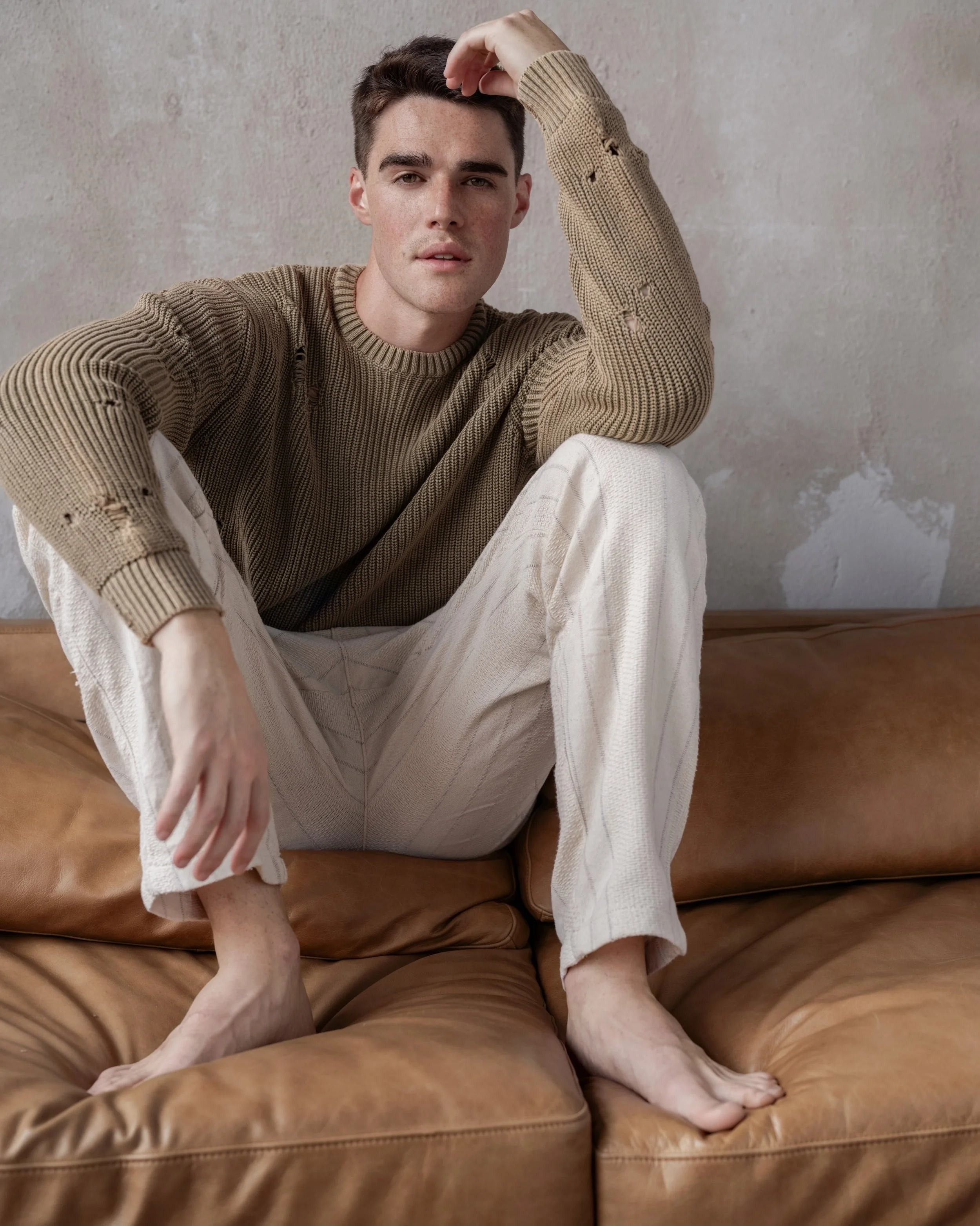 A young man with short brown hair, wearing a distressed beige sweater and cream-colored pants, sitting on a brown leather couch against a textured wall.