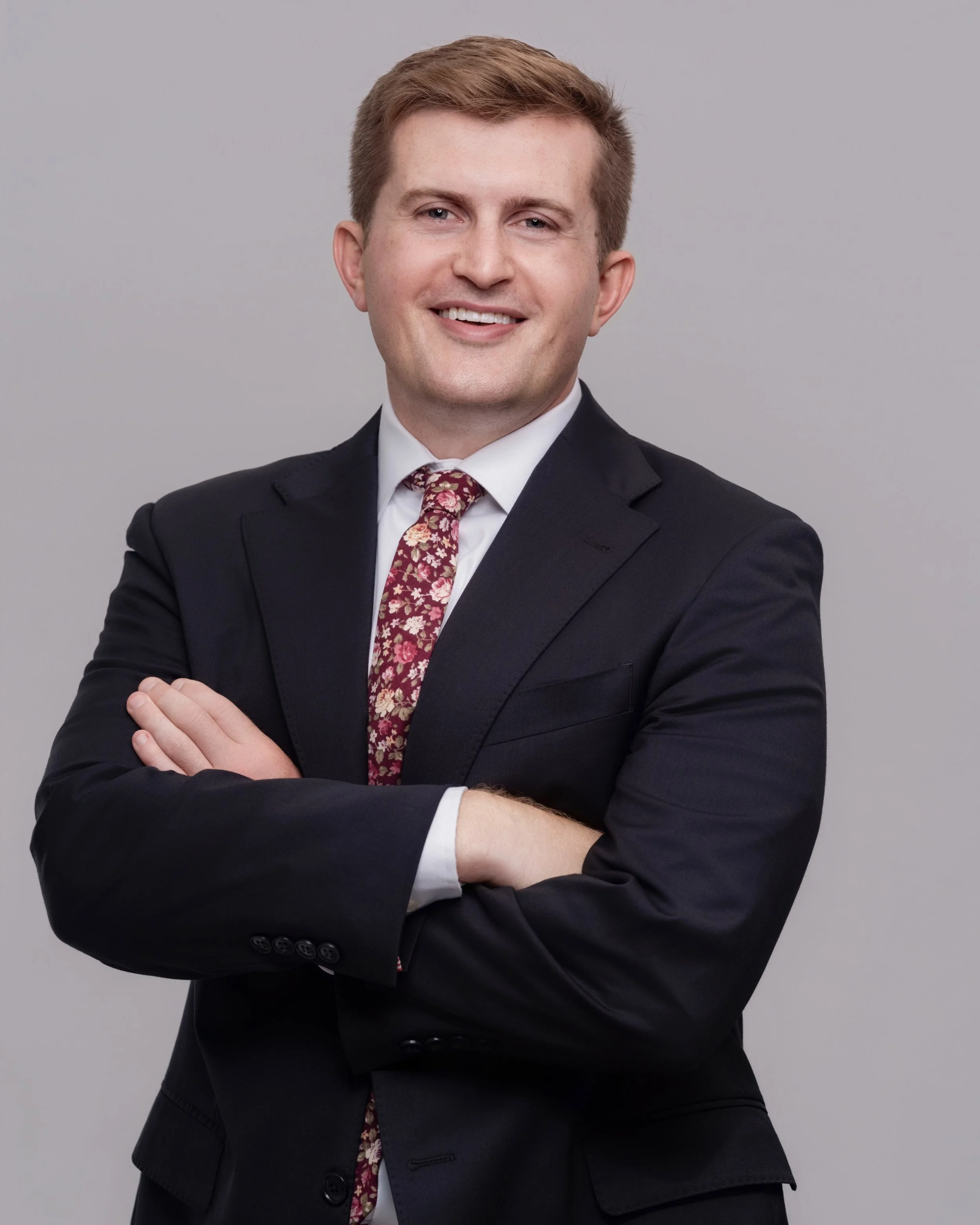 A young man with light brown hair wearing a black suit, white shirt, and a red floral tie, smiling with arms crossed against a gray background.