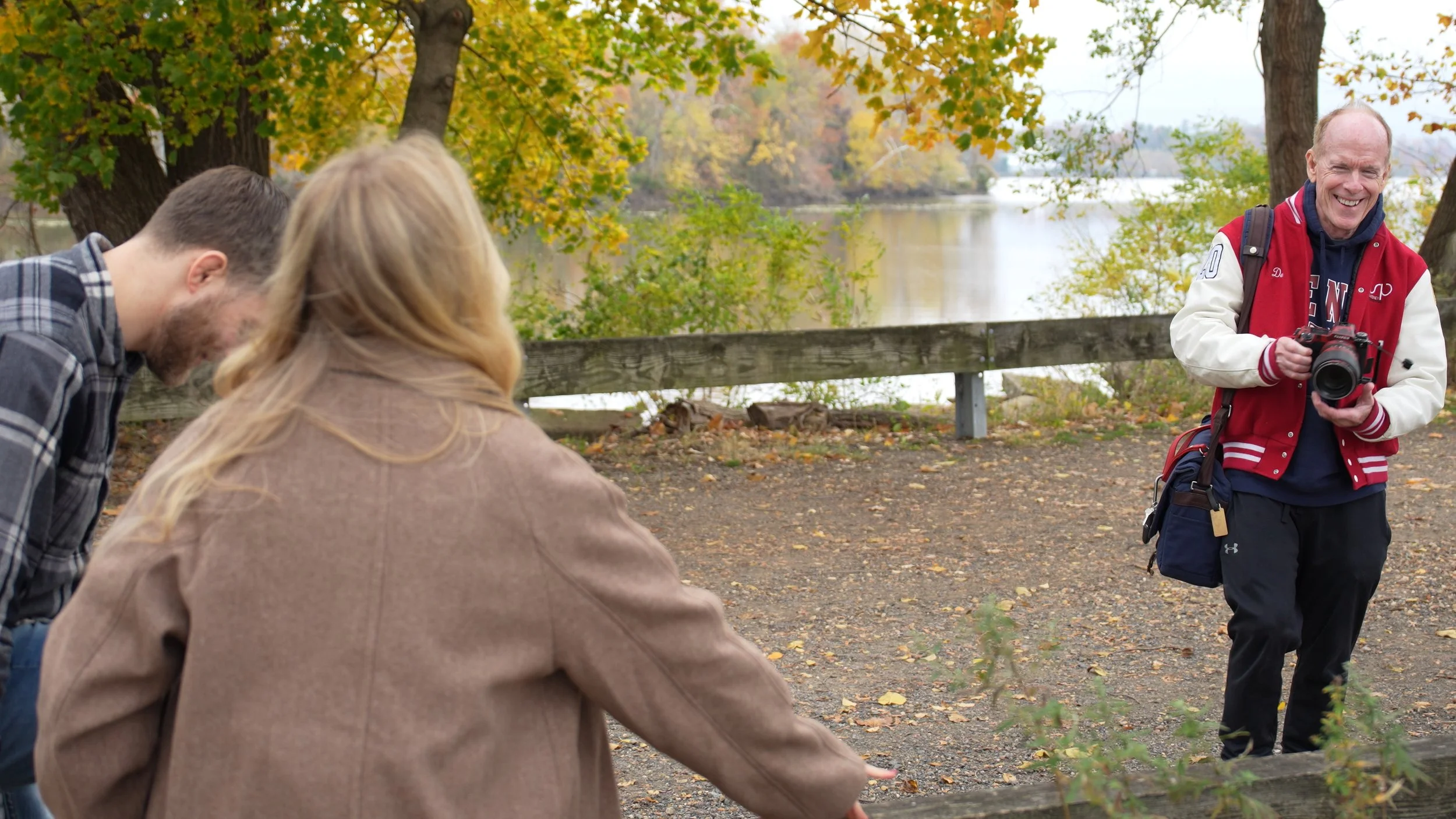 A group of people outdoors near a lake during fall, with trees showing autumn foliage. One person is smiling and holding a camera, another appears to be pointing or reaching towards something, and a third person is observing.