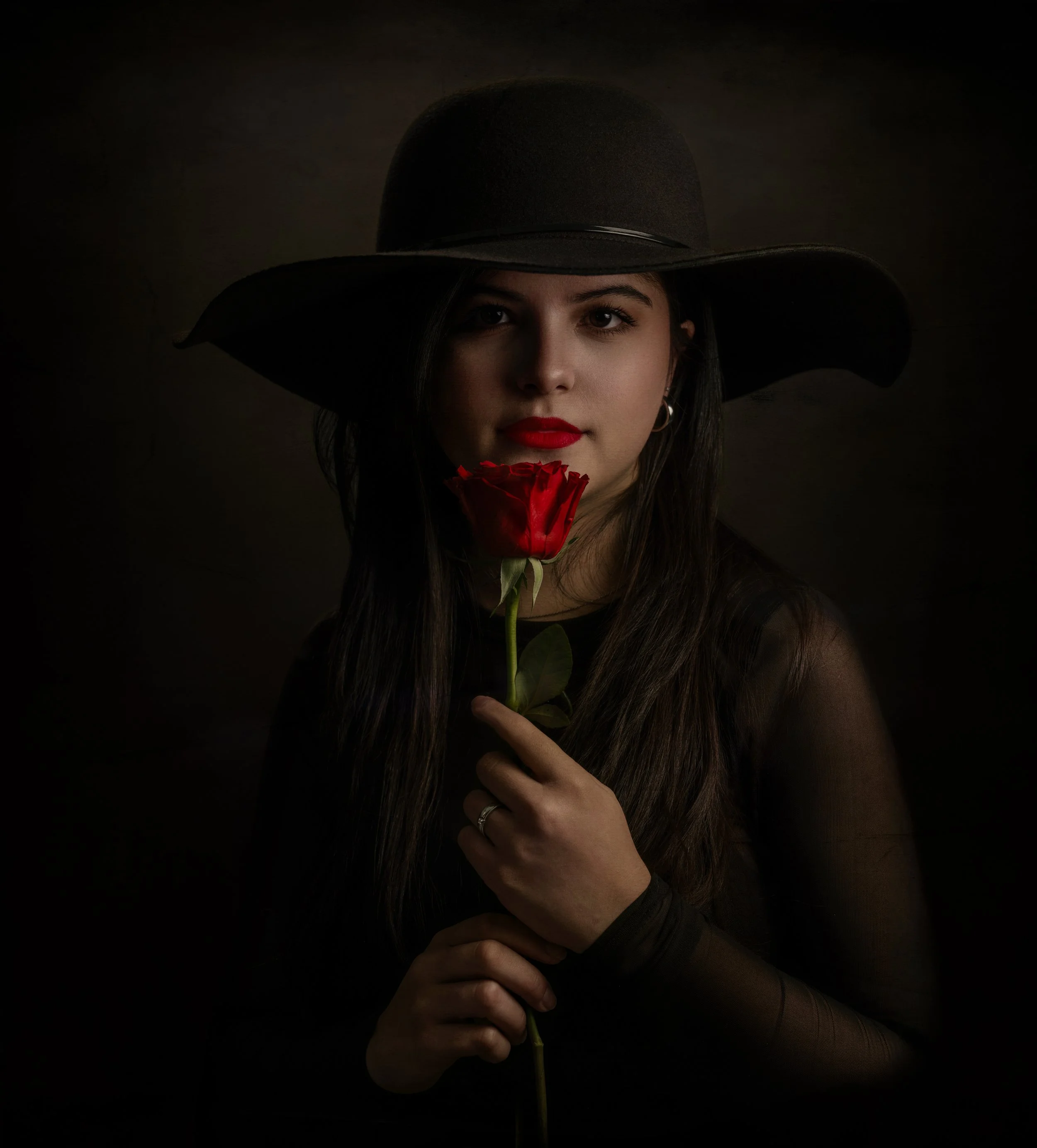 A woman with long dark hair, wearing a large black hat and sheer black top, holding a red rose in front of her face, with only her lips and part of her face visible, against a dark background.