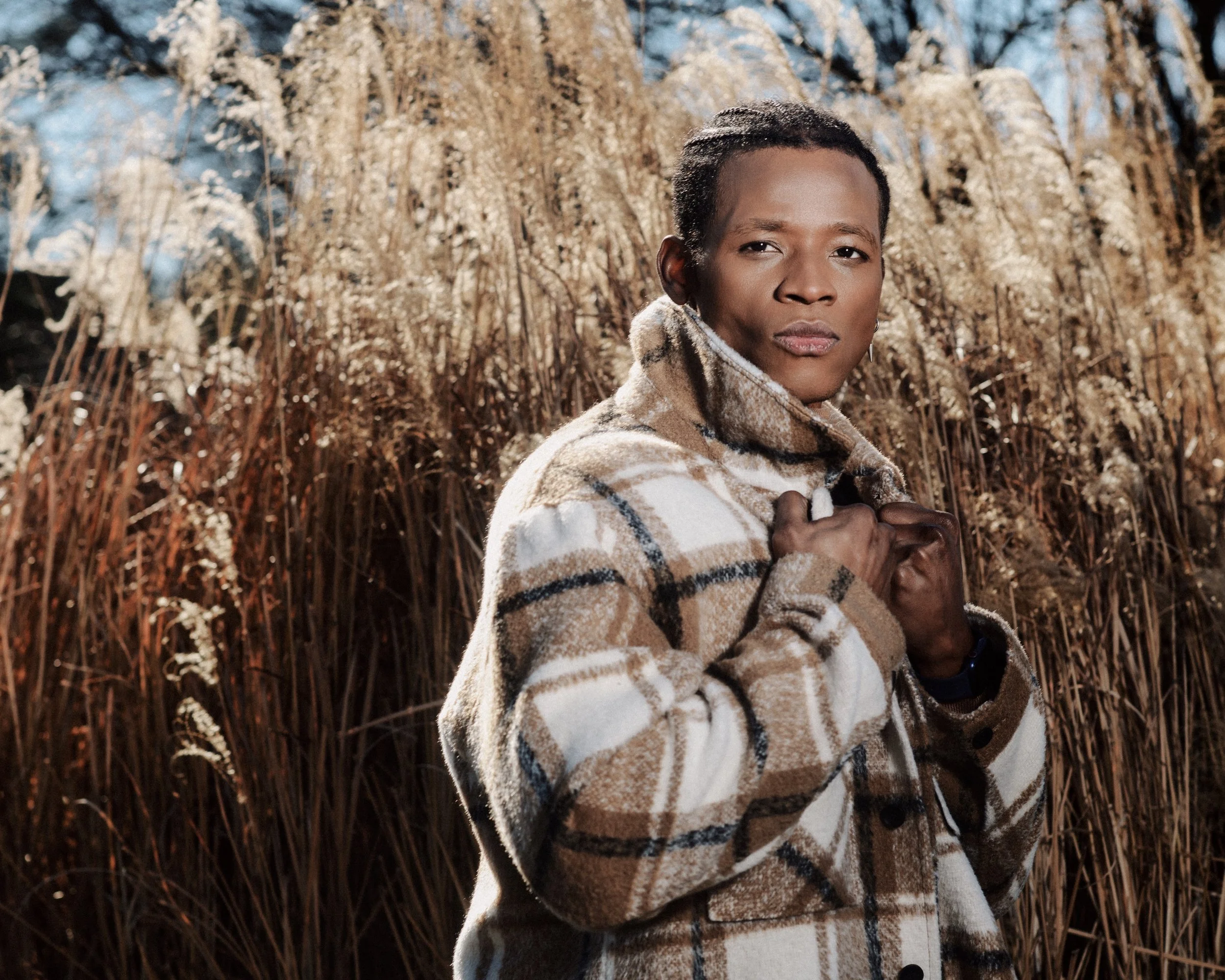 A woman with short hair wearing a beige plaid jacket standing outdoors among tall, dried grasses, holding the collar of her jacket with both hands, looking at the camera.