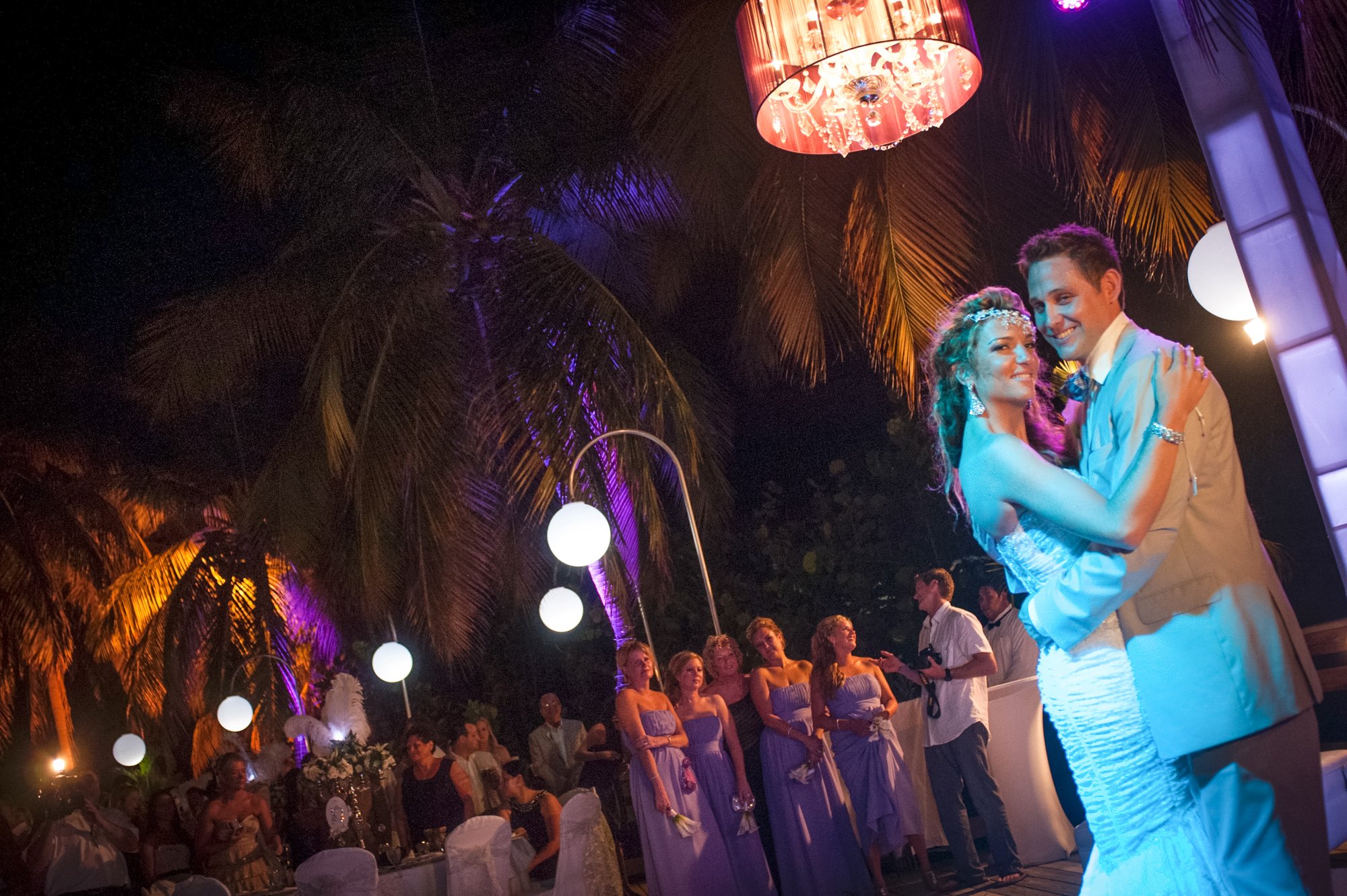 A bride and groom dance closely at their wedding reception outdoors at night. The bride wears a white strapless wedding dress and the groom a light-colored suit. Reception guests, some dressed in purple bridesmaid dresses, watch nearby. The scene is 