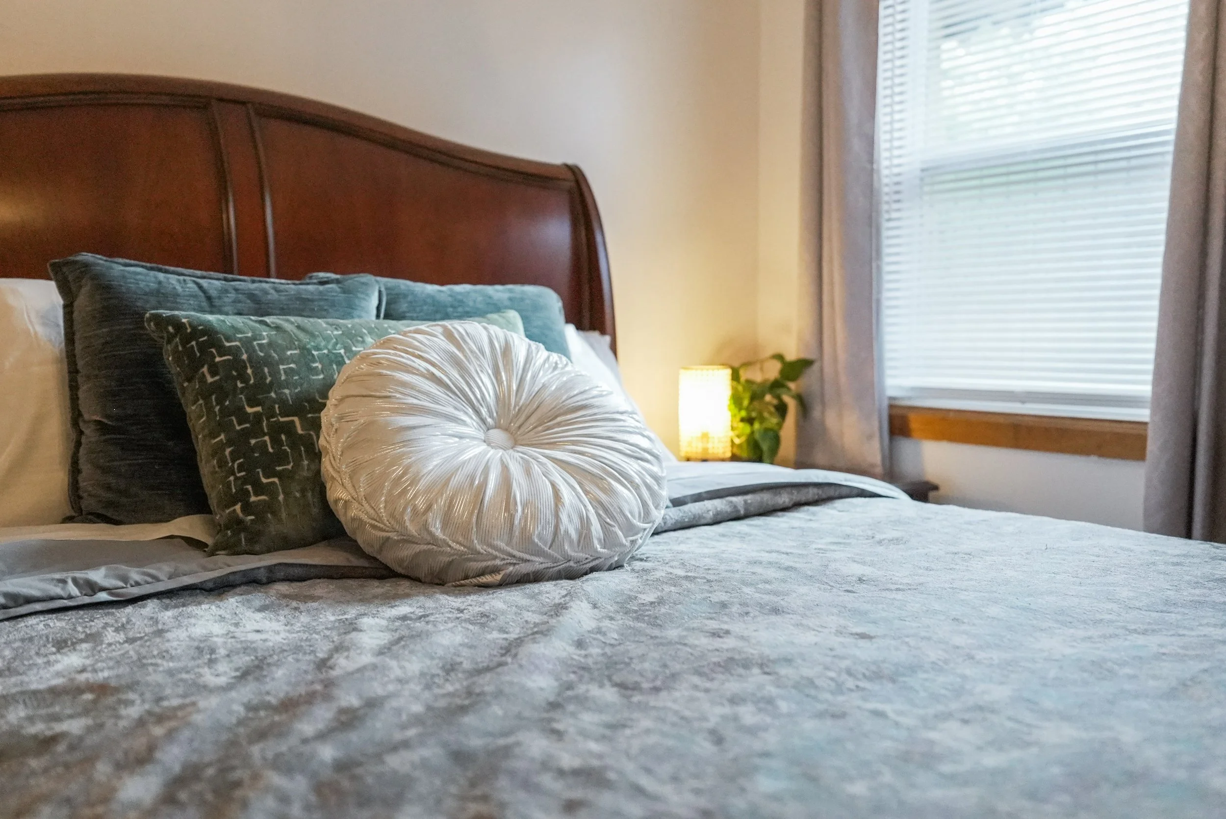 A neatly made bed with multiple pillows, a bedside lamp, and a window with blinds in a cozy bedroom.