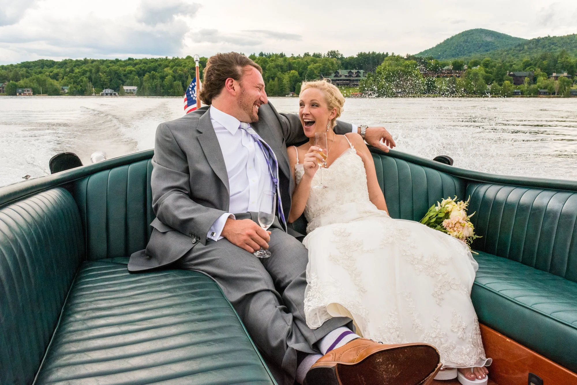 A newlywed couple in wedding attire enjoying champagne on a boat with a scenic lake and mountains in the background.