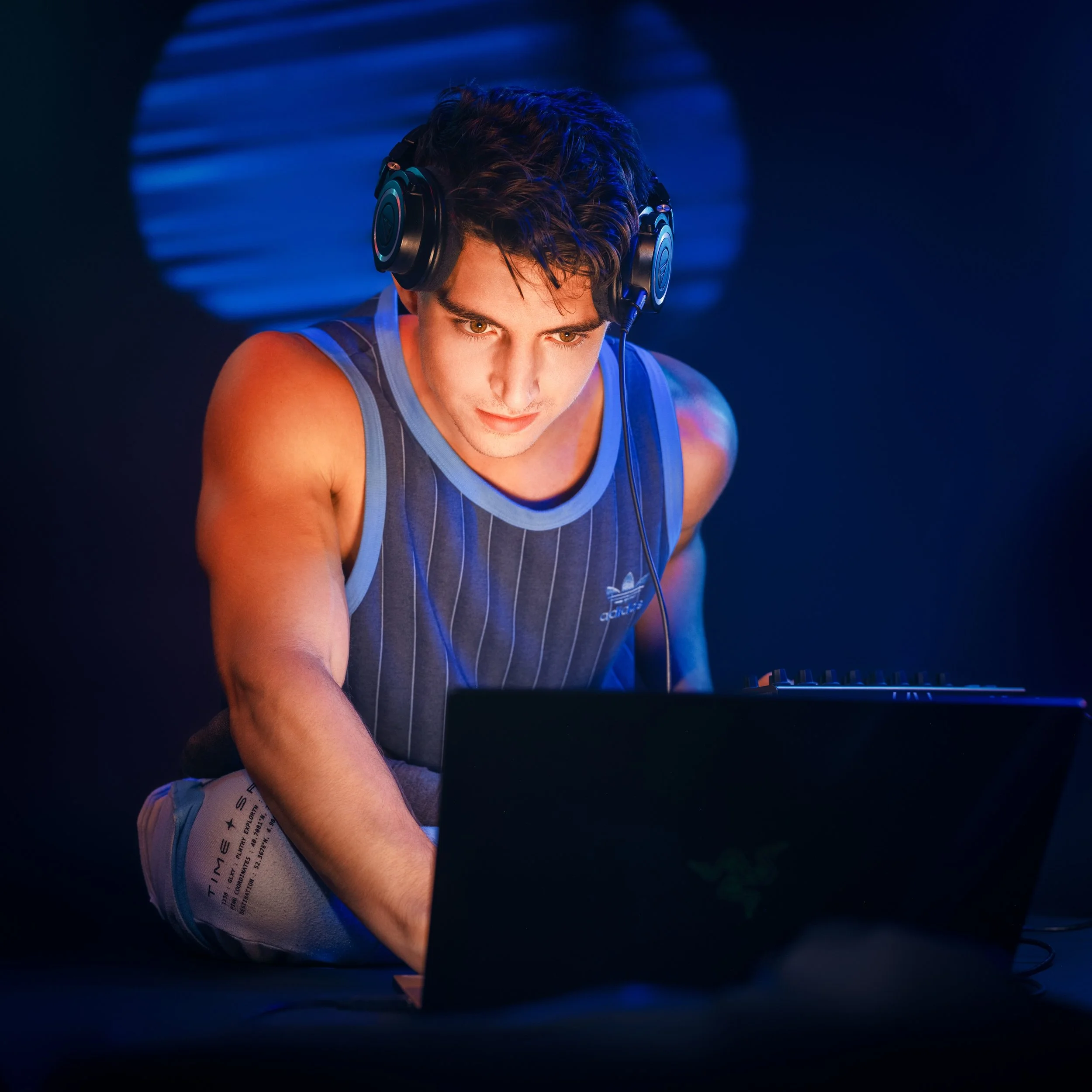 Young man wearing headphones and a sleeveless striped shirt working on a laptop in a dark room with blue lighting.