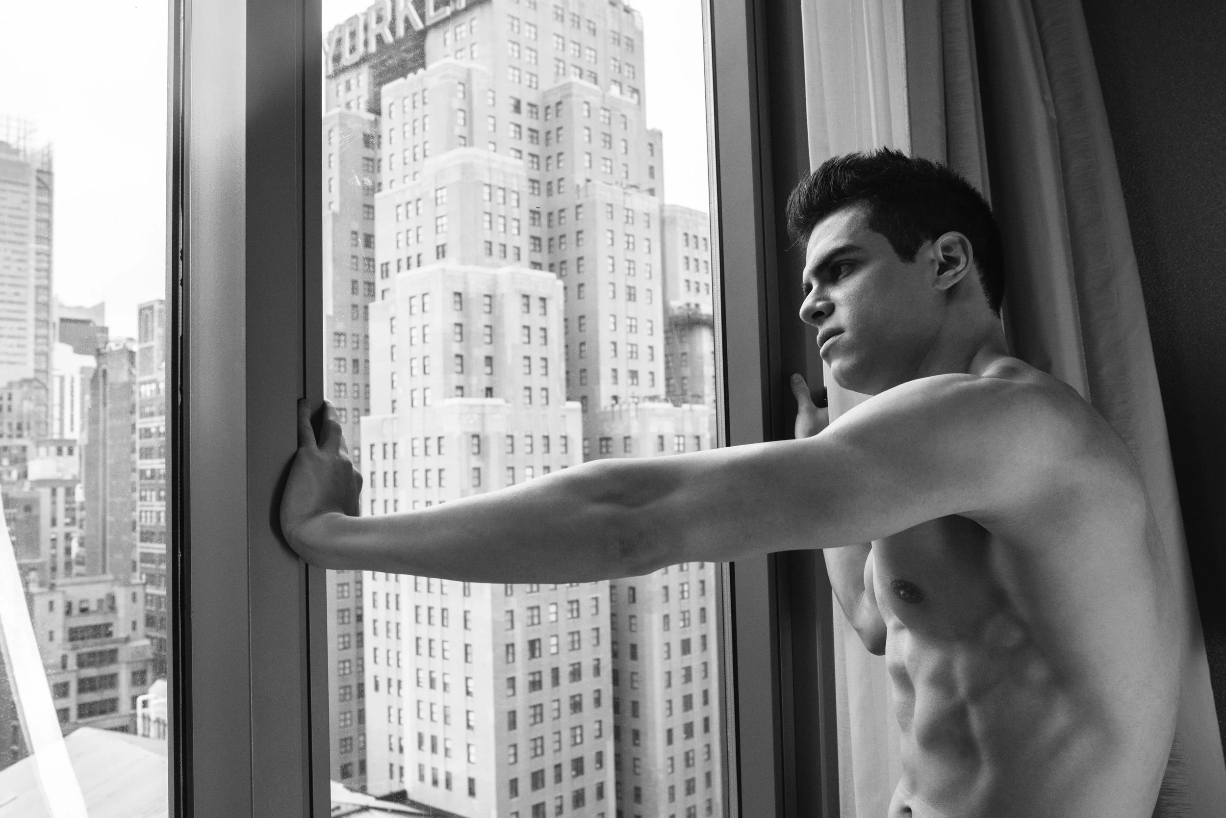 A shirtless young man with dark hair looking contemplative while holding onto a window frame, with city skyscrapers visible outside.