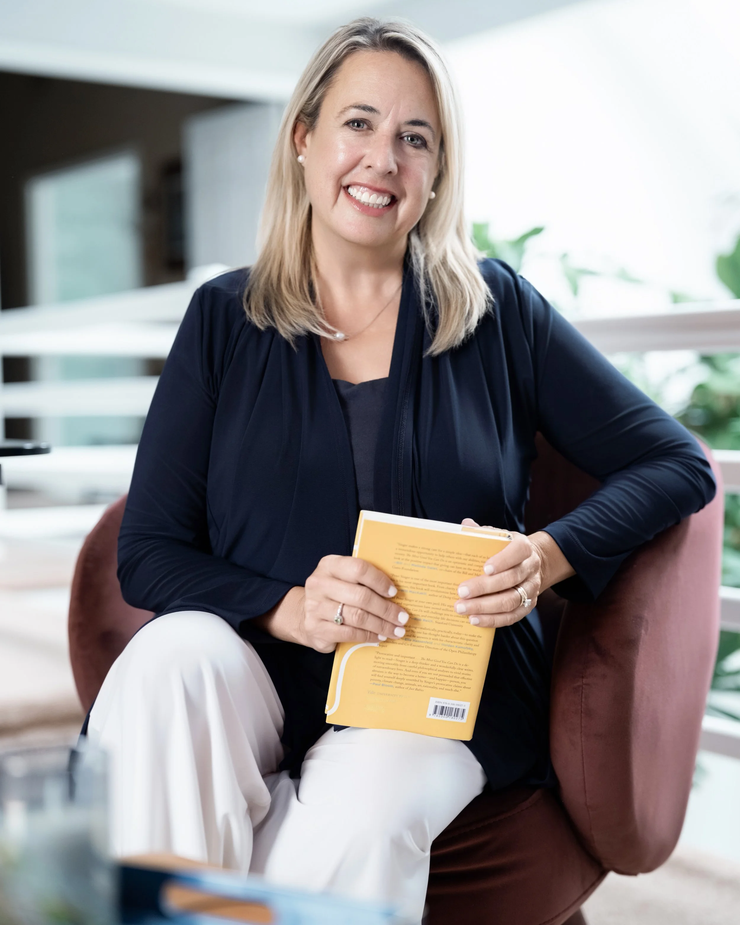 A woman with blonde hair, wearing a navy blue top and white pants, sitting on a pink armchair, holding a yellow book, smiling at the camera in a bright indoor space with plants in the background.