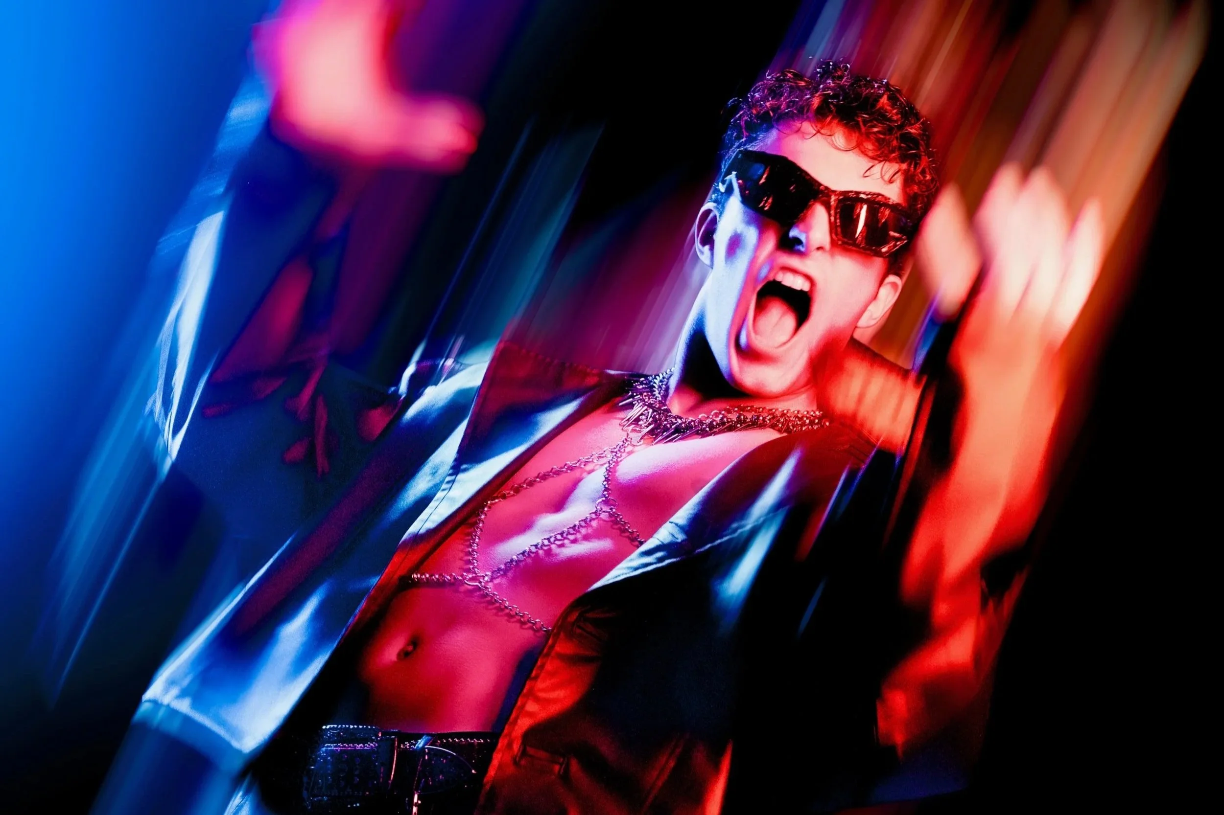 A person with curly hair wearing dark sunglasses and a leather jacket, shouting with their mouth open, surrounded by colorful, neon-like lighting.