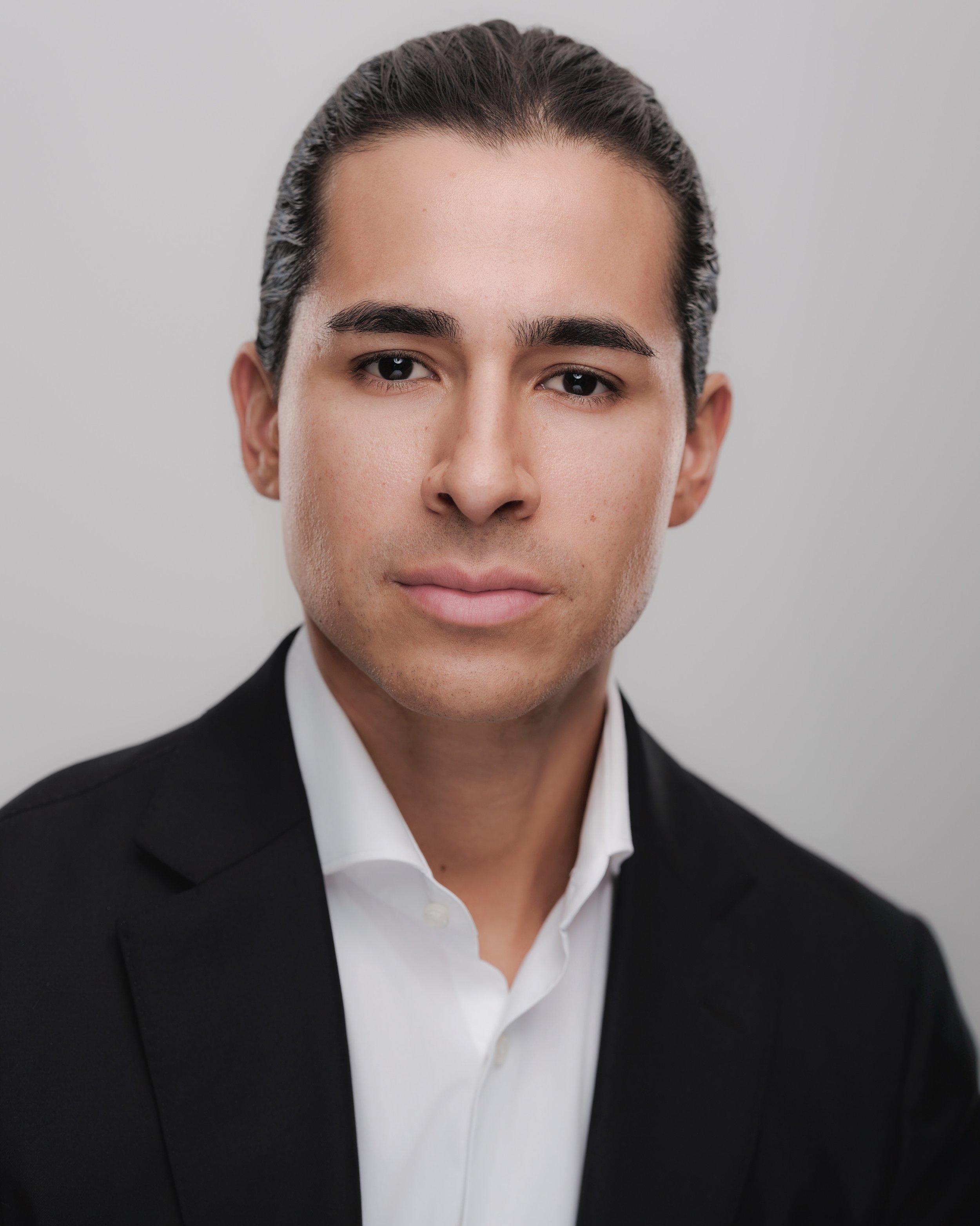 Professional headshot of a man with dark hair tied back, wearing a black suit and white shirt, against a light gray background.