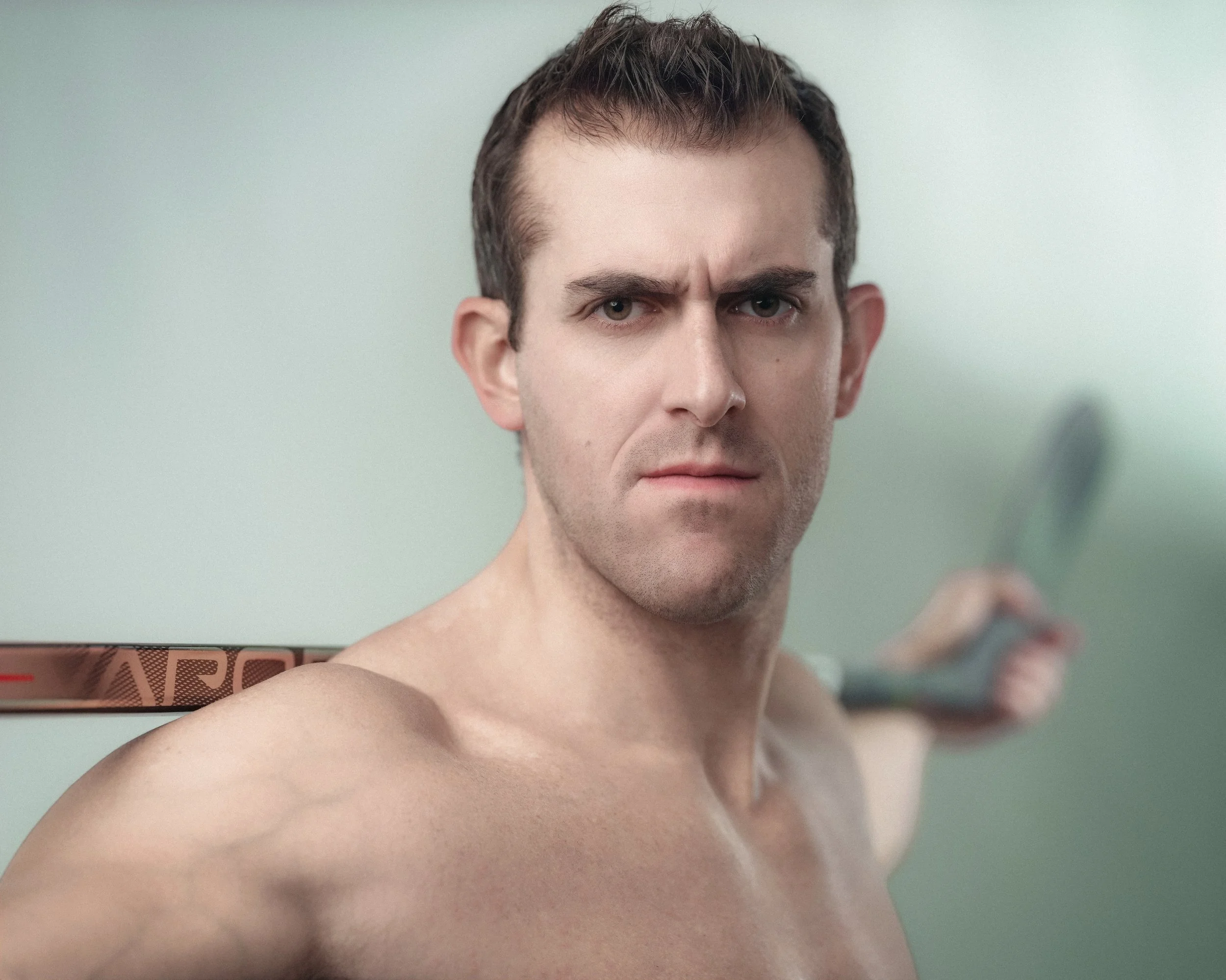 A shirtless man with short brown hair, furrowed brow, and a serious expression holding a hockey stick behind his head, looking directly at the camera against a plain, light-colored background.